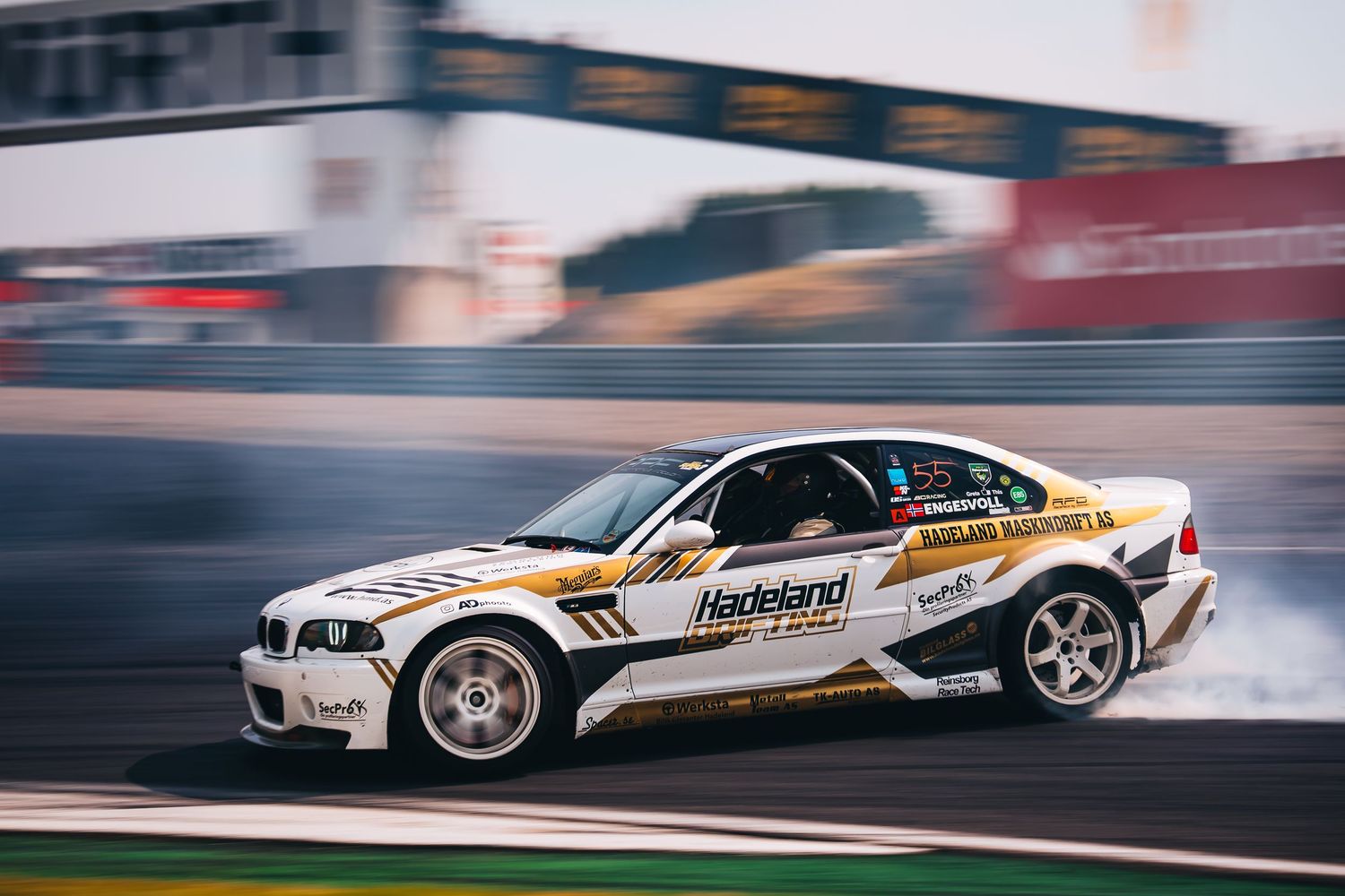 A white BMW drift car with racing livery slides sideways on a racetrack during a competition.