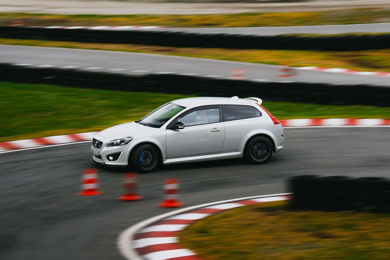 White sports car racing around a track with red and white curbing and orange traffic cones visible.