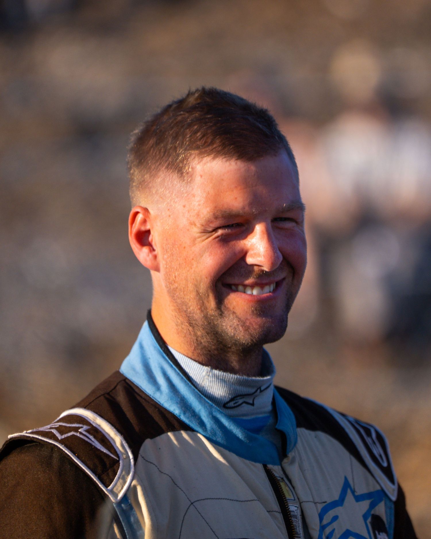 A smiling person in racing gear wearing a blue and black suit illuminated by golden sunlight.