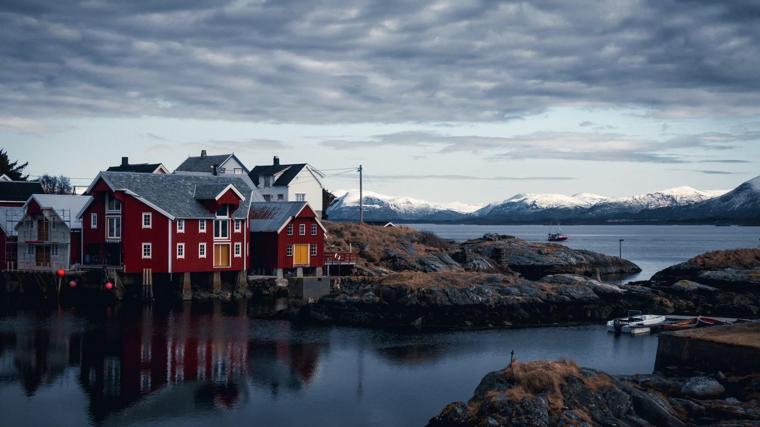 Red wooden houses reflect on calm waters against dramatic cloudy sky and mountain backdrop in Norwegian fishing village.