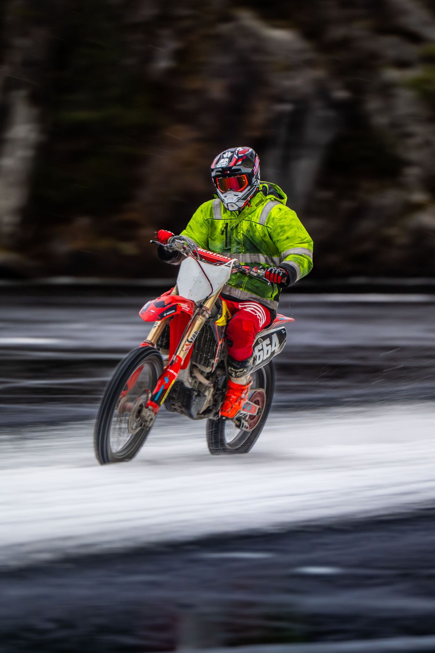 A motorcyclist in bright yellow gear rides a red dirt bike across a snowy winter landscape with motion blur effect.