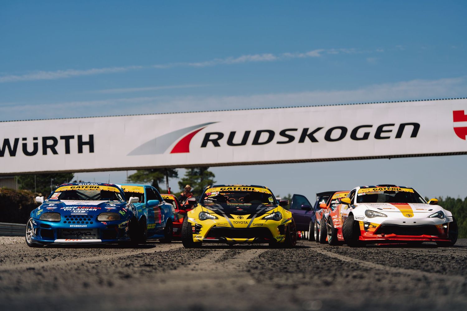 Colorful racing cars lined up on a track against a blue sky with dramatic lighting and shadows.