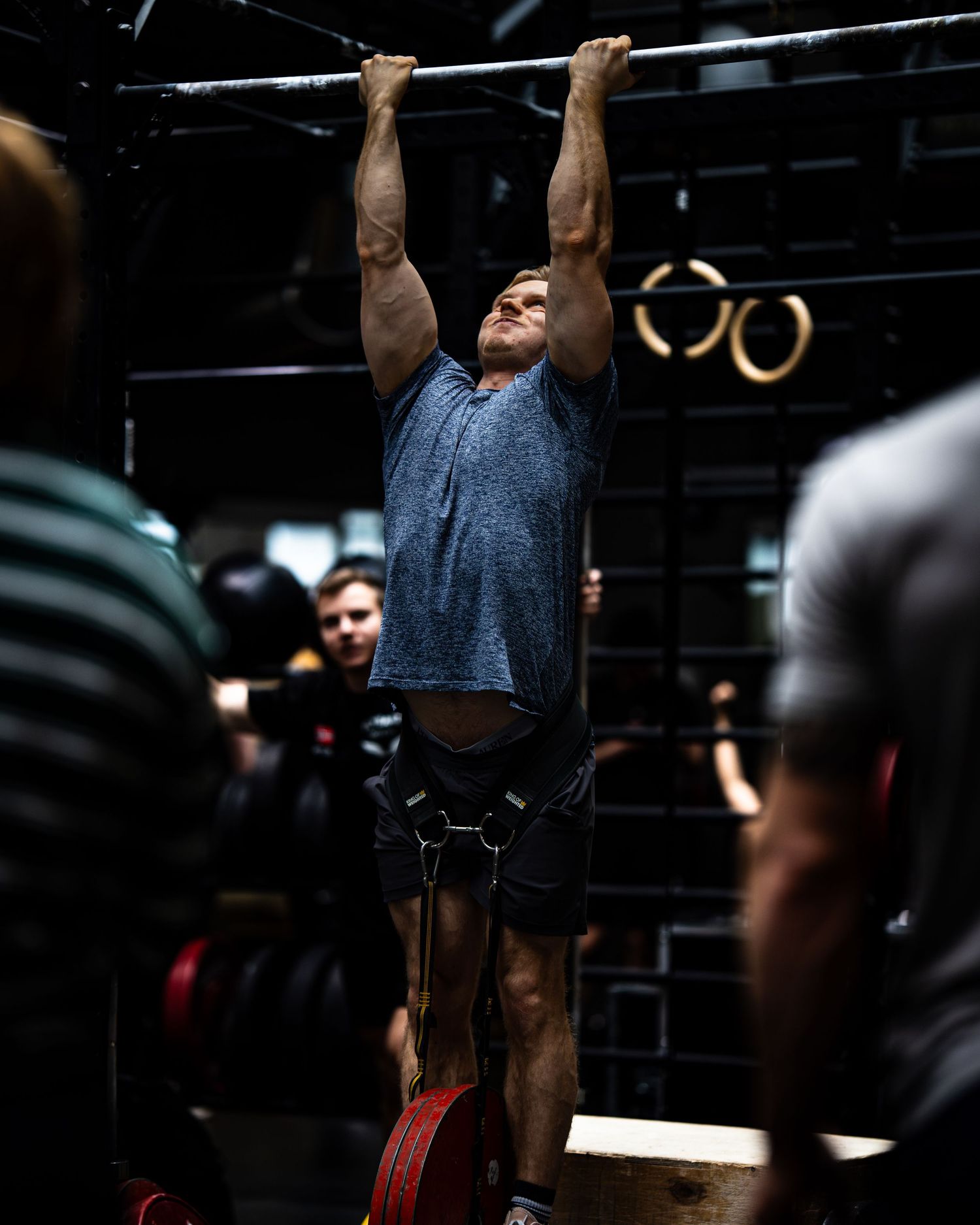 Muscular athlete performs weighted pull-up in indoor gym with gymnastics rings visible in background.