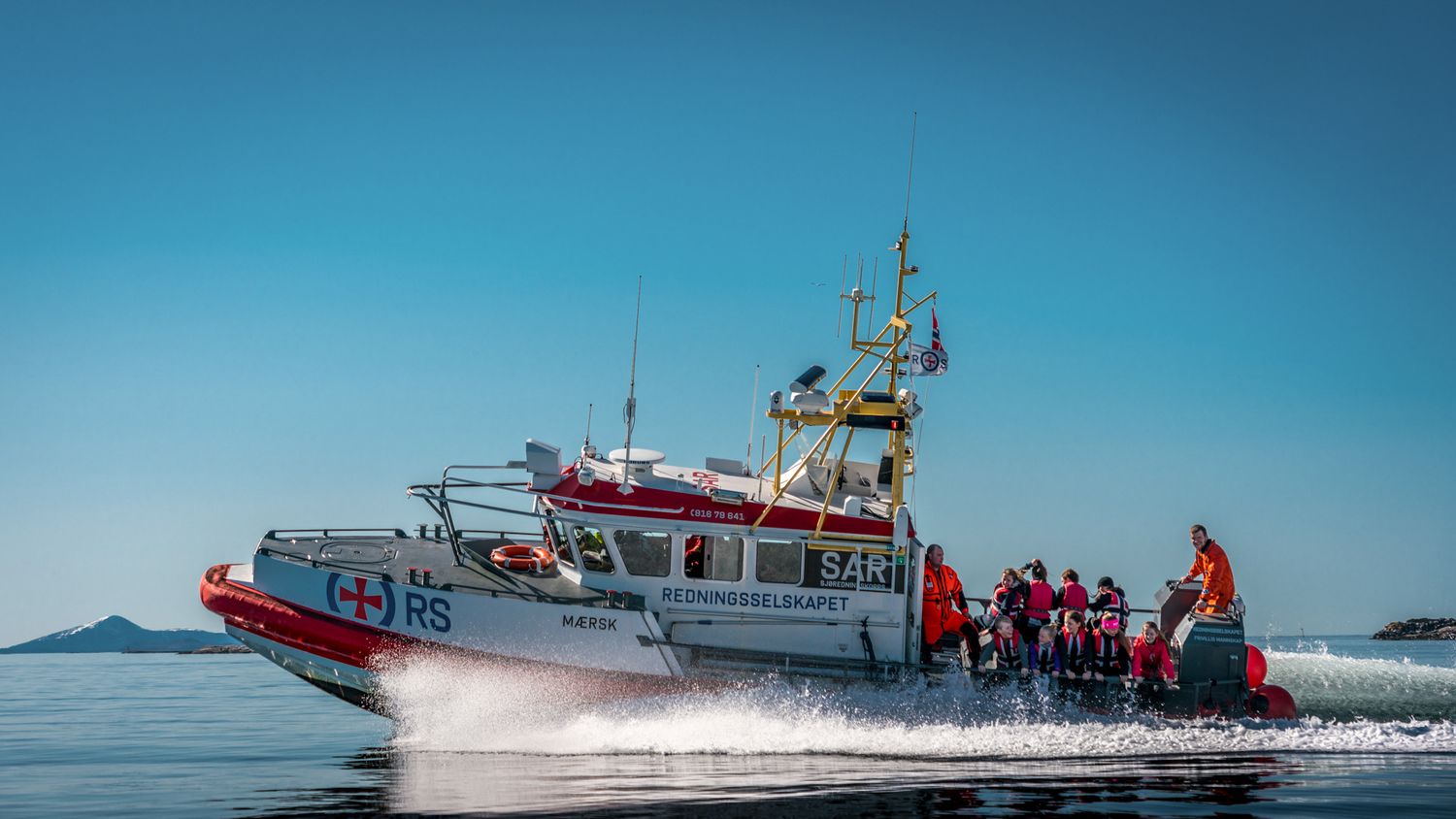 Red and white rescue vessel speeding across blue waters with mountains in background.