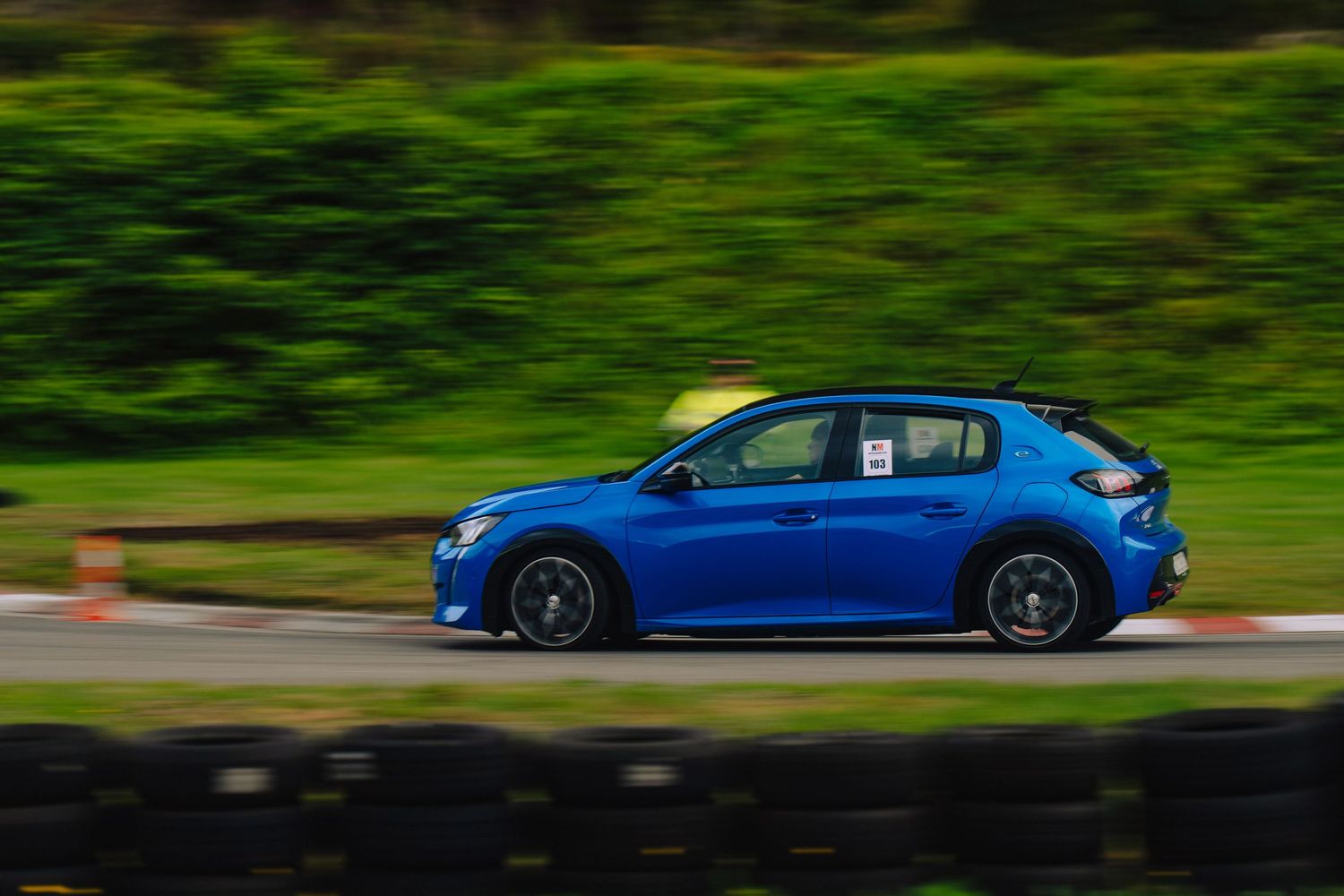 A blue sports car drives along a winding road with lush green foliage in the background.