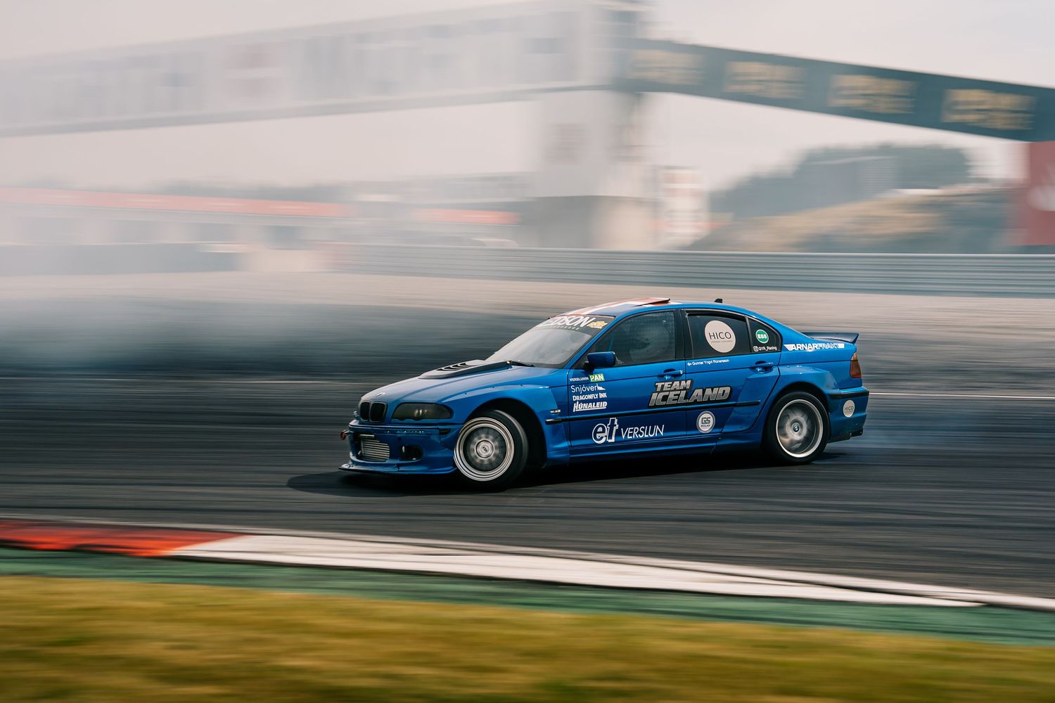 Blue drift car slides sideways on racetrack creating smoke during competitive motorsport event.