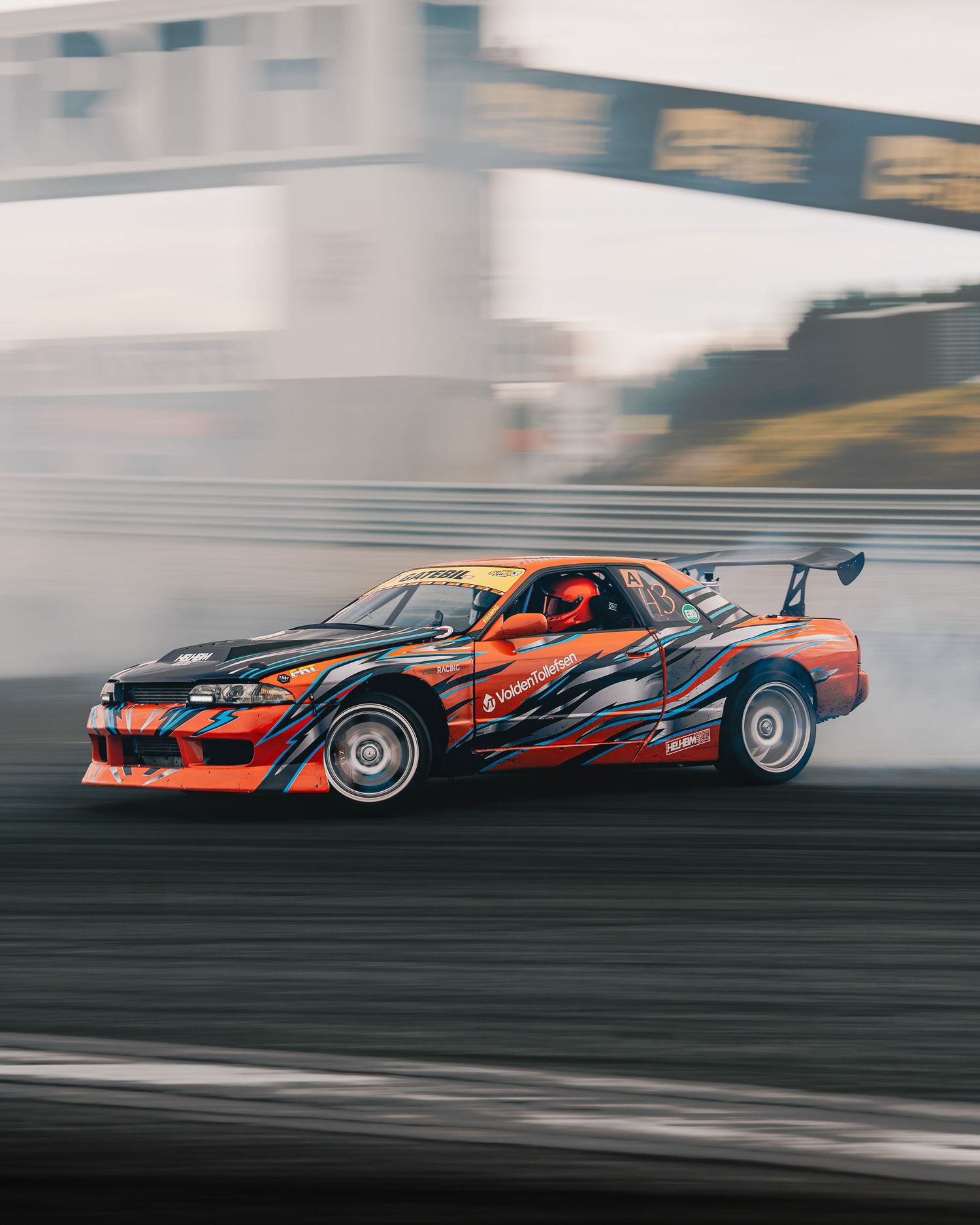 Orange drift car slides sideways creating smoke while drifting on race track during competition.
