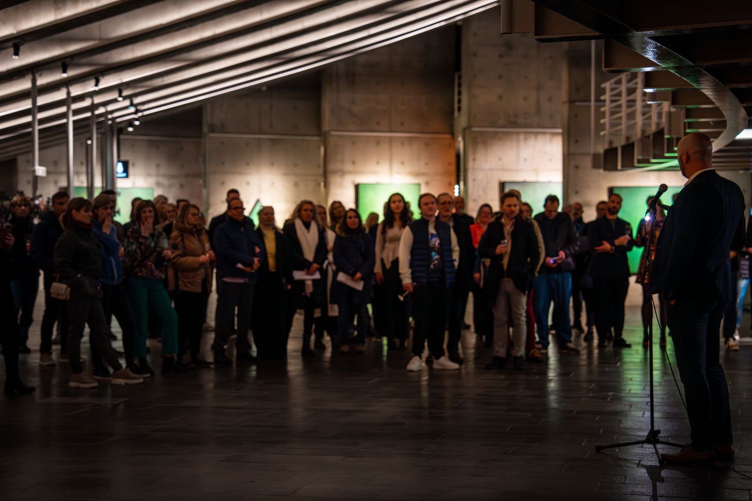 Art exhibition attendees listen to a presentation in a modern industrial space.