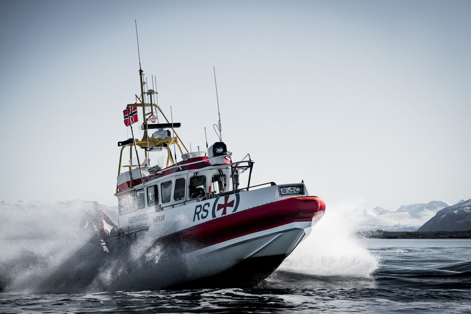 Maritime rescue boat cutting through stormy seas with snow-covered peaks in distance.