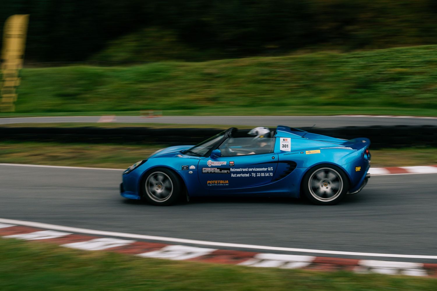 Racing car speeds through a race track corner with green trees in the background.