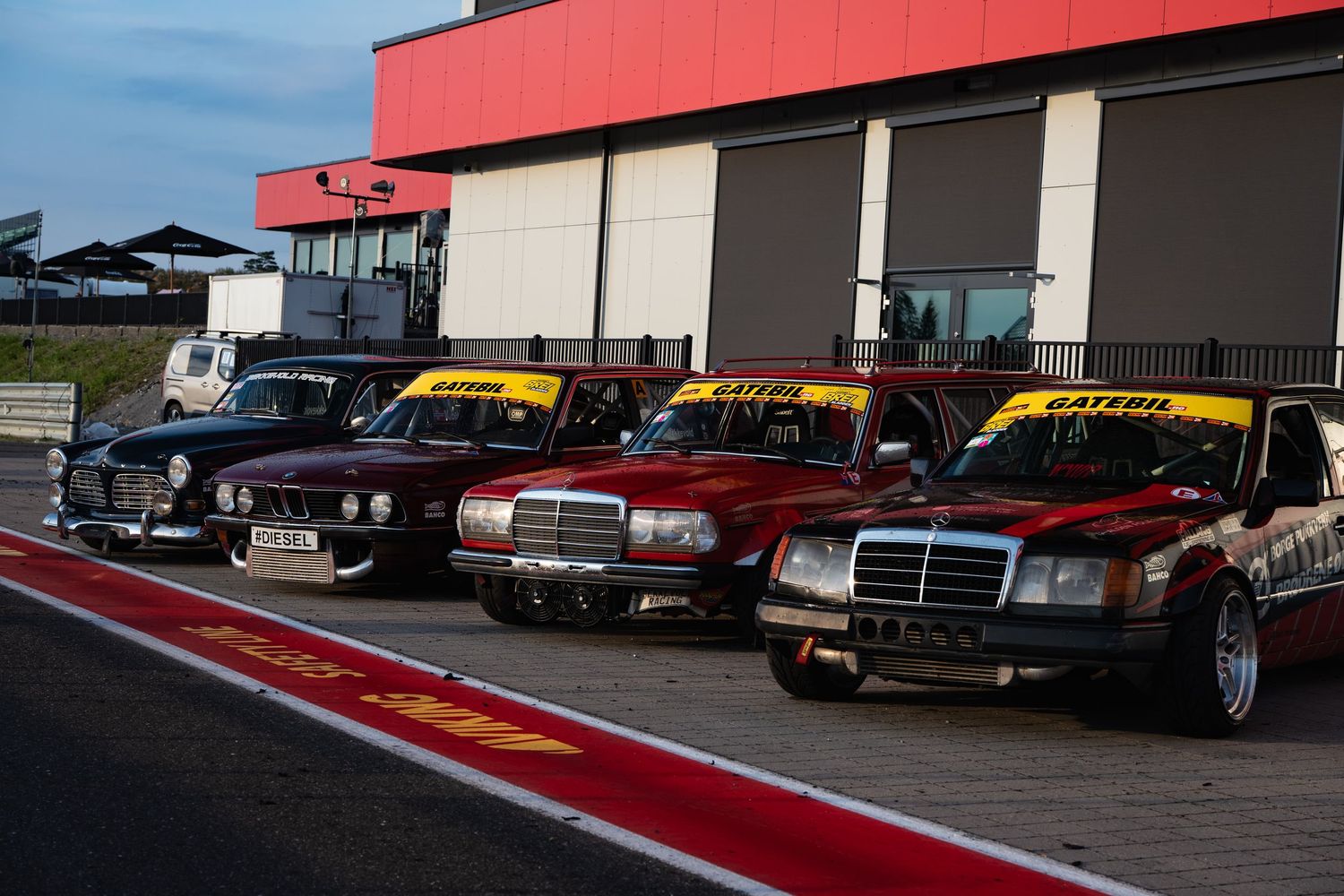 Row of classic Mercedes-Benz sedans lined up at a race track with red and black exterior features.