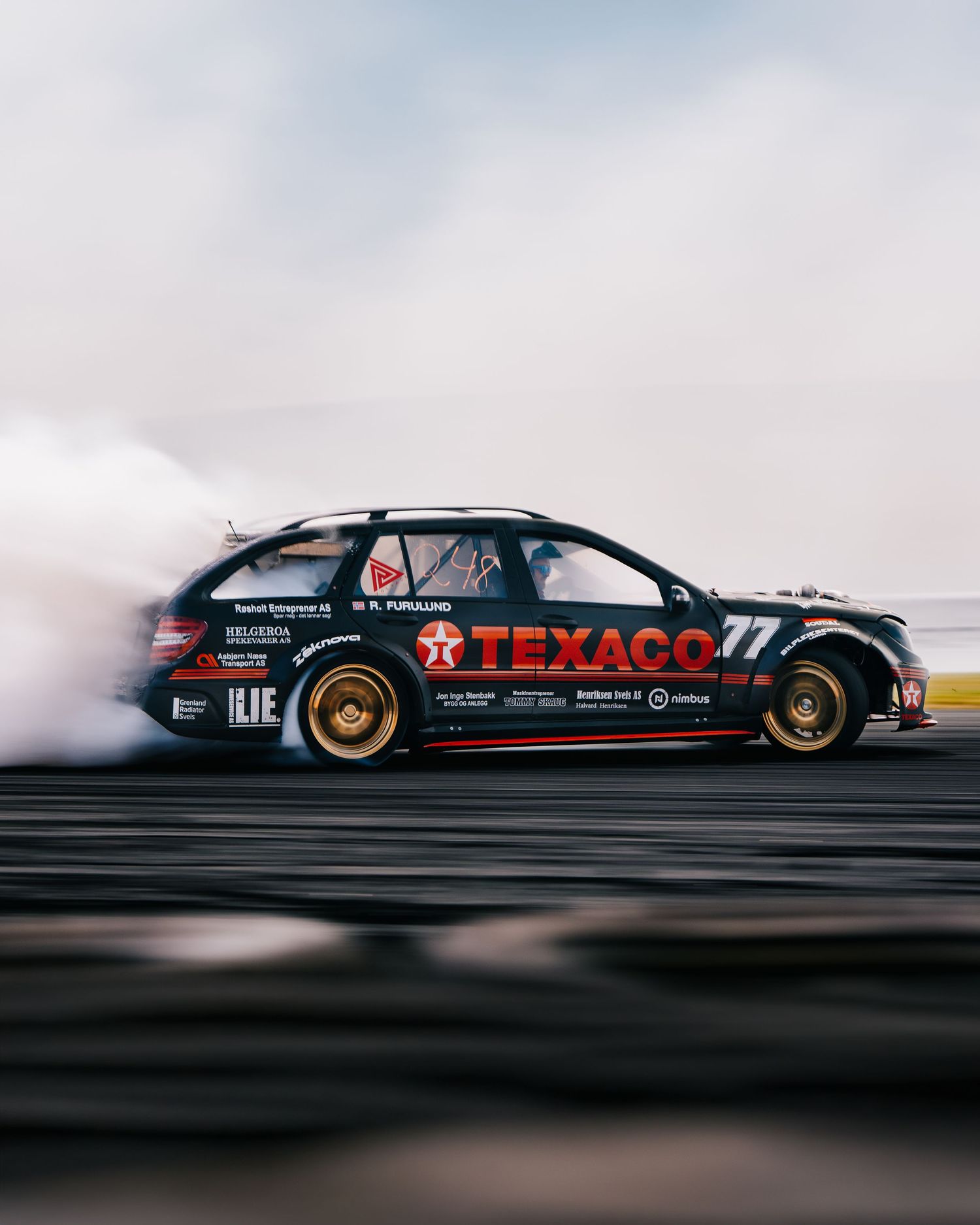 A black race car with Texaco branding drifts sideways on a track during a dynamic motorsport performance.