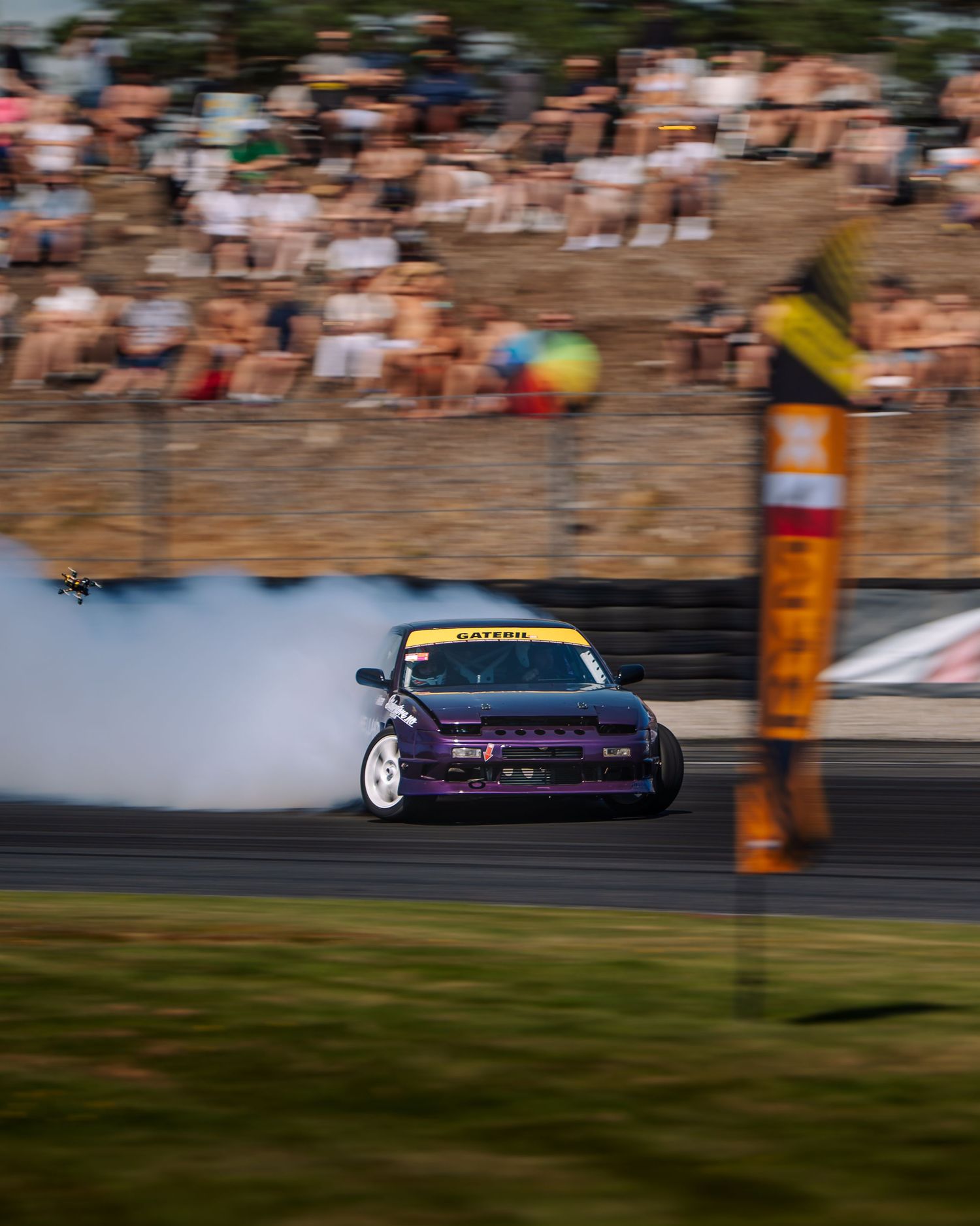 A blurred motion shot of a purple sports car drifting on a racetrack with spectators visible in the background.