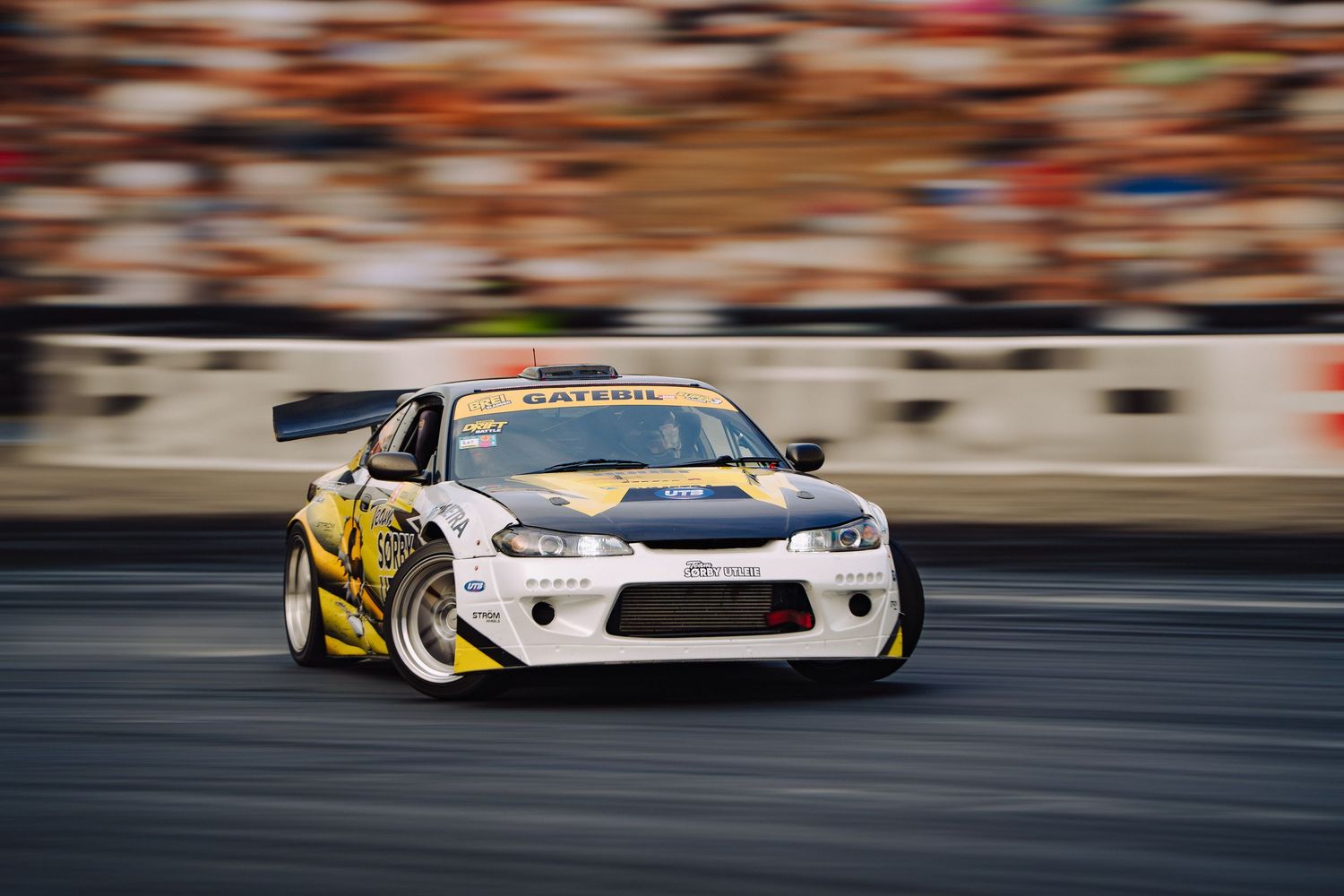 Racing cars speed past spectators during a motorsport competition with motion blur effect capturing their velocity.