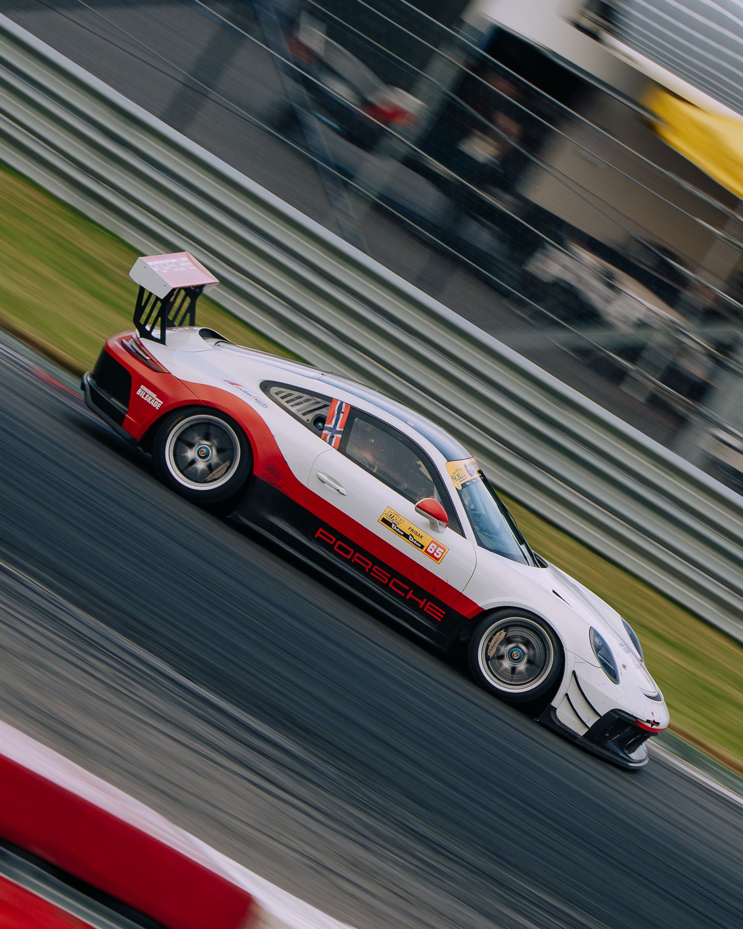 A white and red Porsche race car speeds along a racetrack with motion blur and grandstands visible in the background.