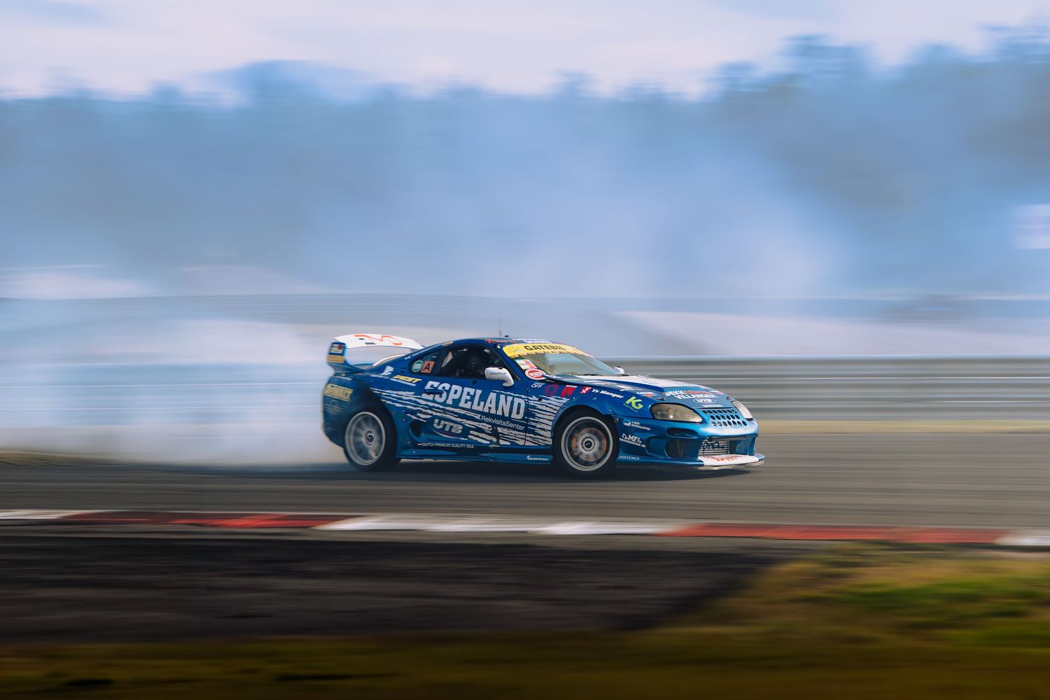 A blue racing car speeds around a racetrack with motion blur against a cloudy sky background.