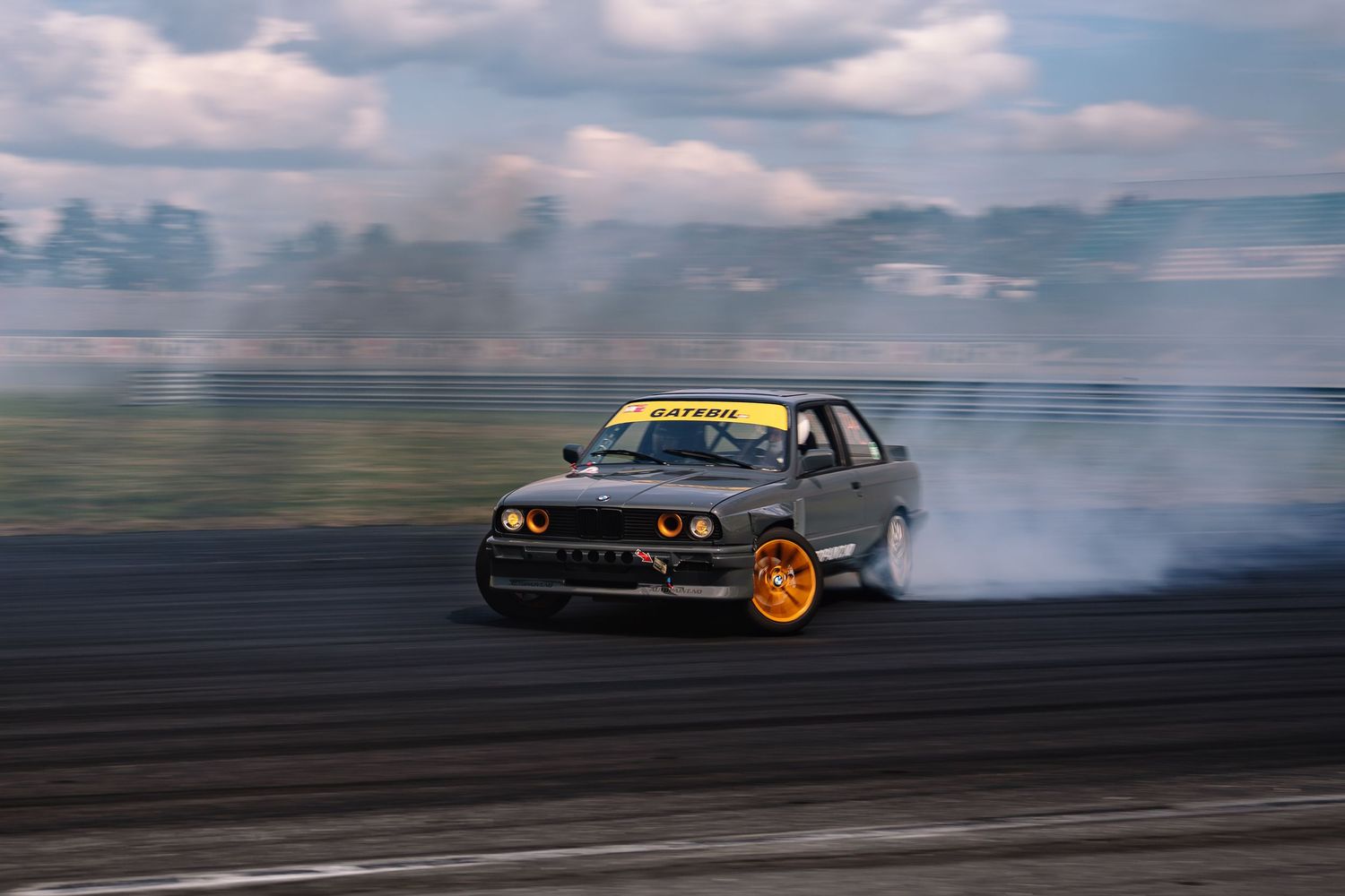 A BMW M3 drifts sideways on a racetrack with smoke billowing from its orange wheels during a drift competition.