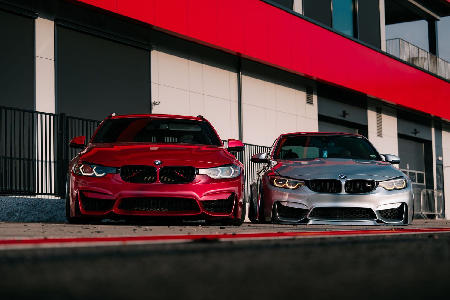 Two BMW M-series sports cars, one red and one silver, parked side by side in front of a modern building.