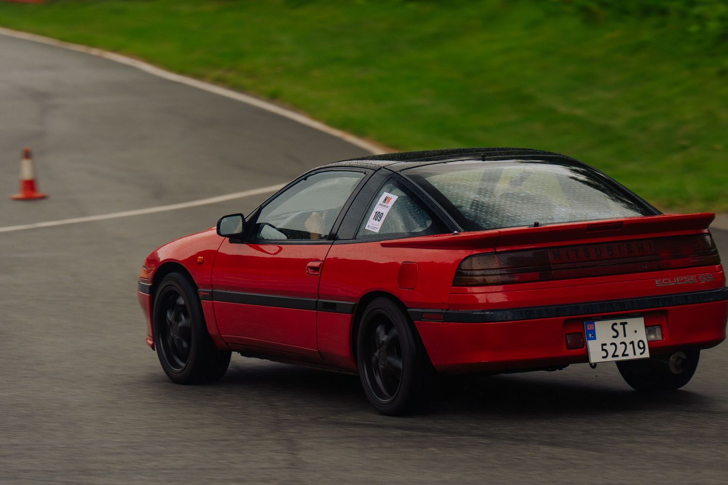 A red sports car drives on a wet racetrack with lush green surroundings in the background.