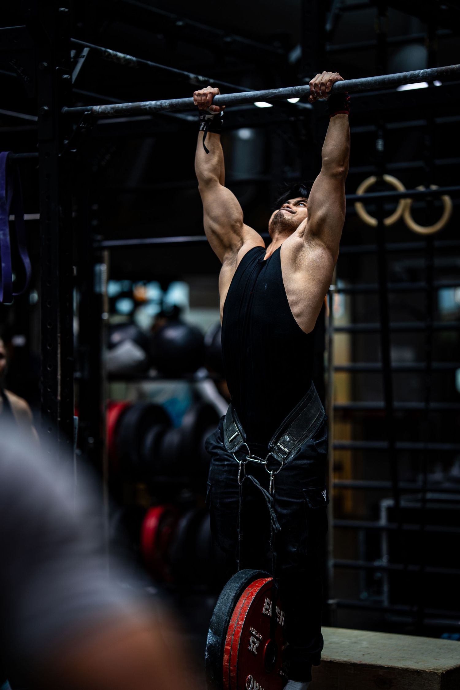 Athlete executes weighted pull-up exercise with intense focus in dimly lit fitness facility.