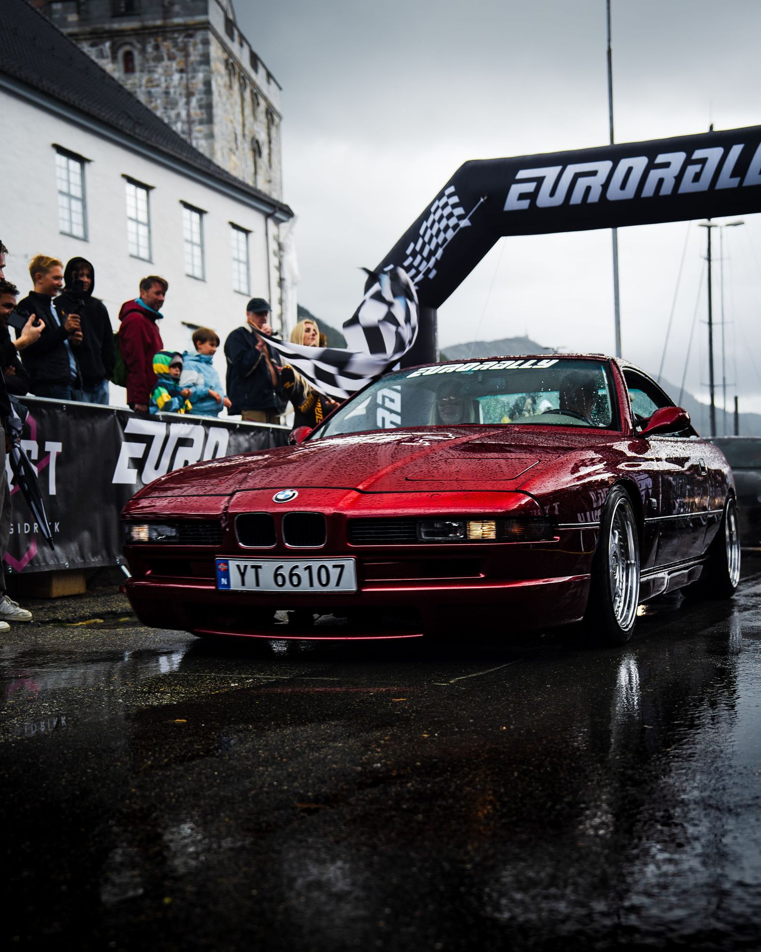 Red BMW 8 Series classic car passing under Eurorally arch during rainy event.