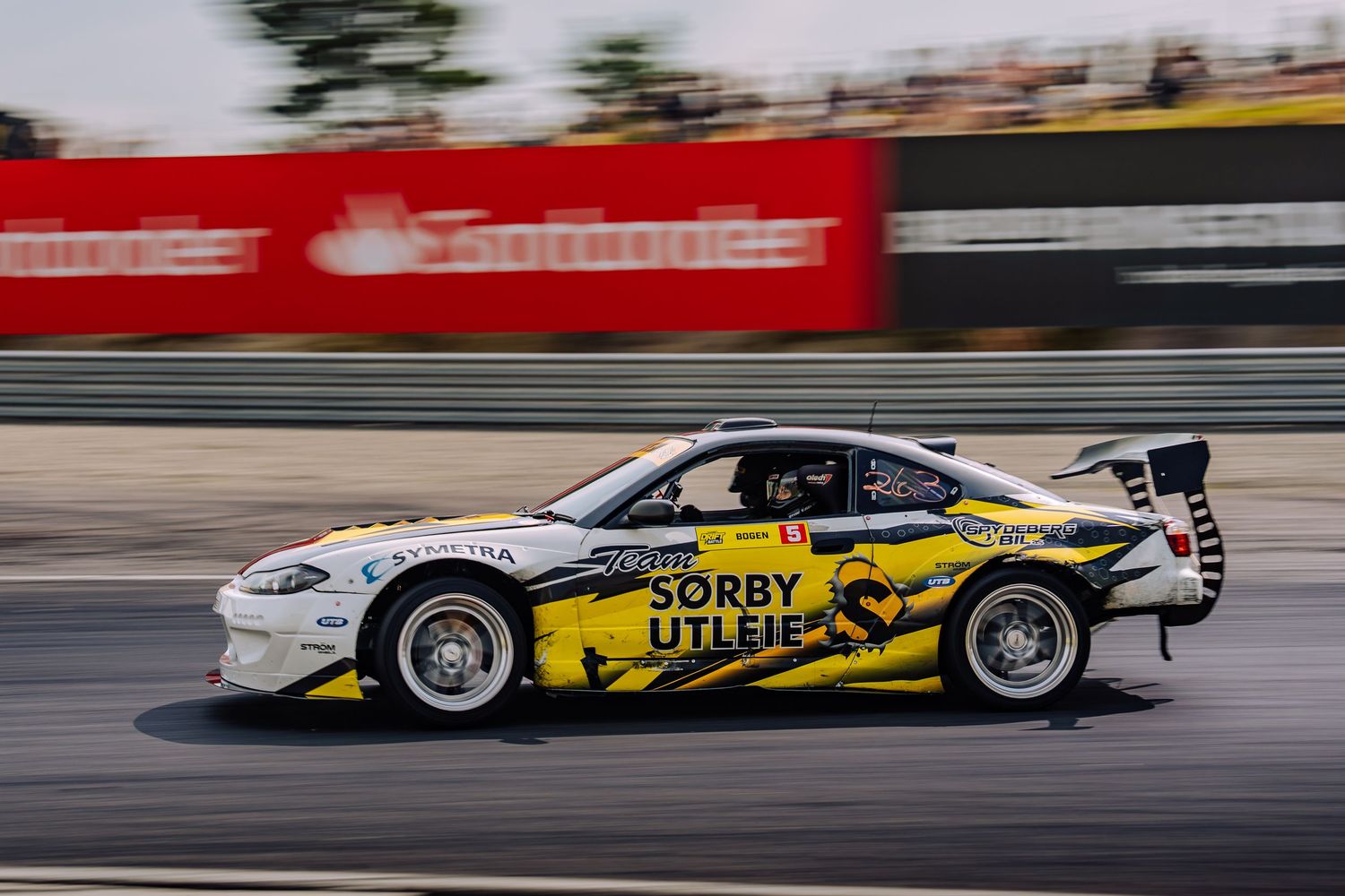 Race cars speed past on a track showing motion blur with yellow and white livery designs on display.