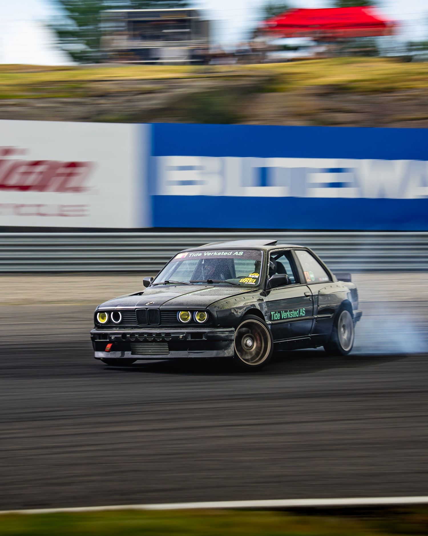 A BMW E30 drifting on a race track with smoke coming from the tires during a motorsport competition.