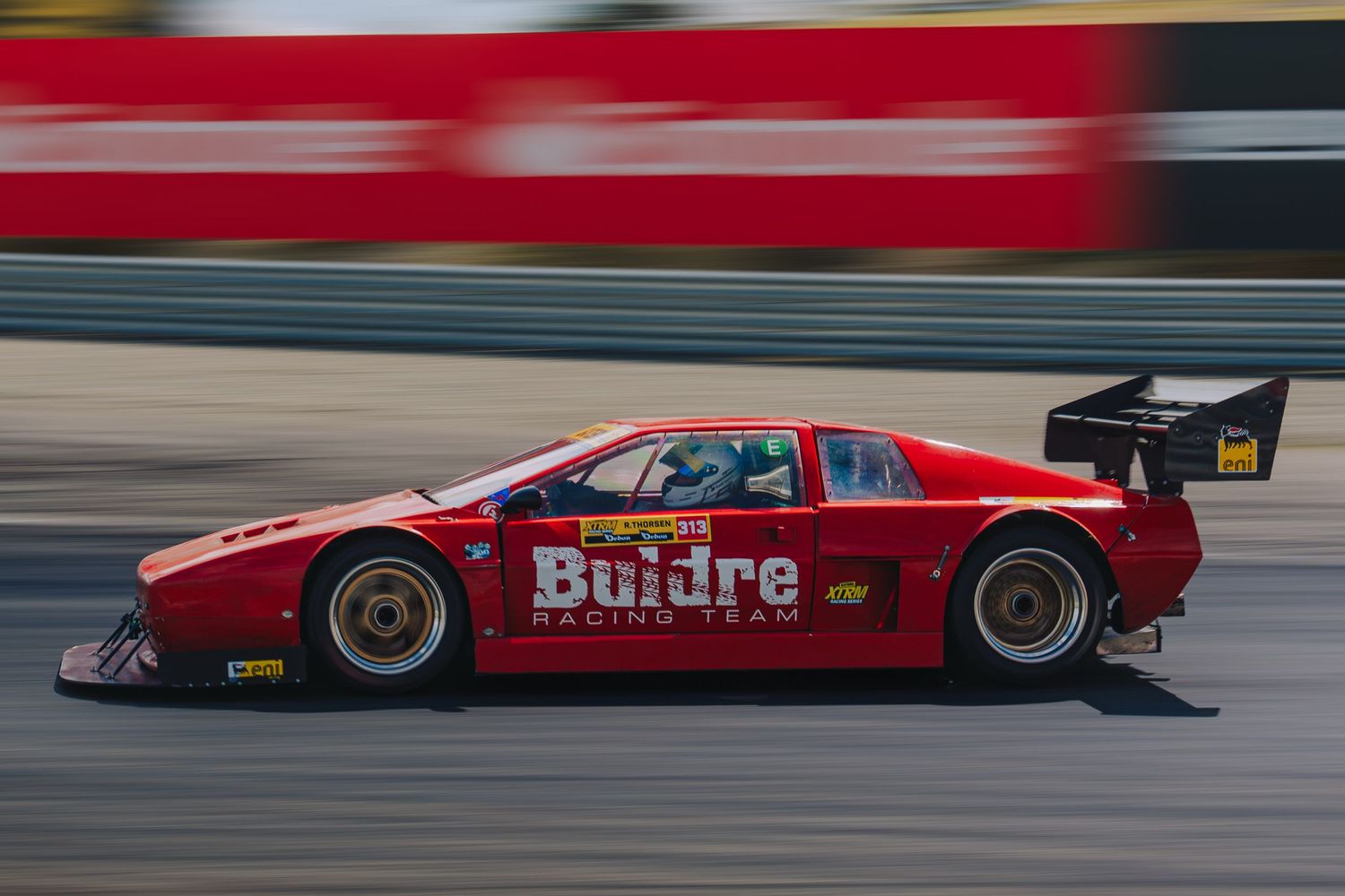 Red racing car with Buidre sponsorship speeds along racetrack with motion blur effect in background.