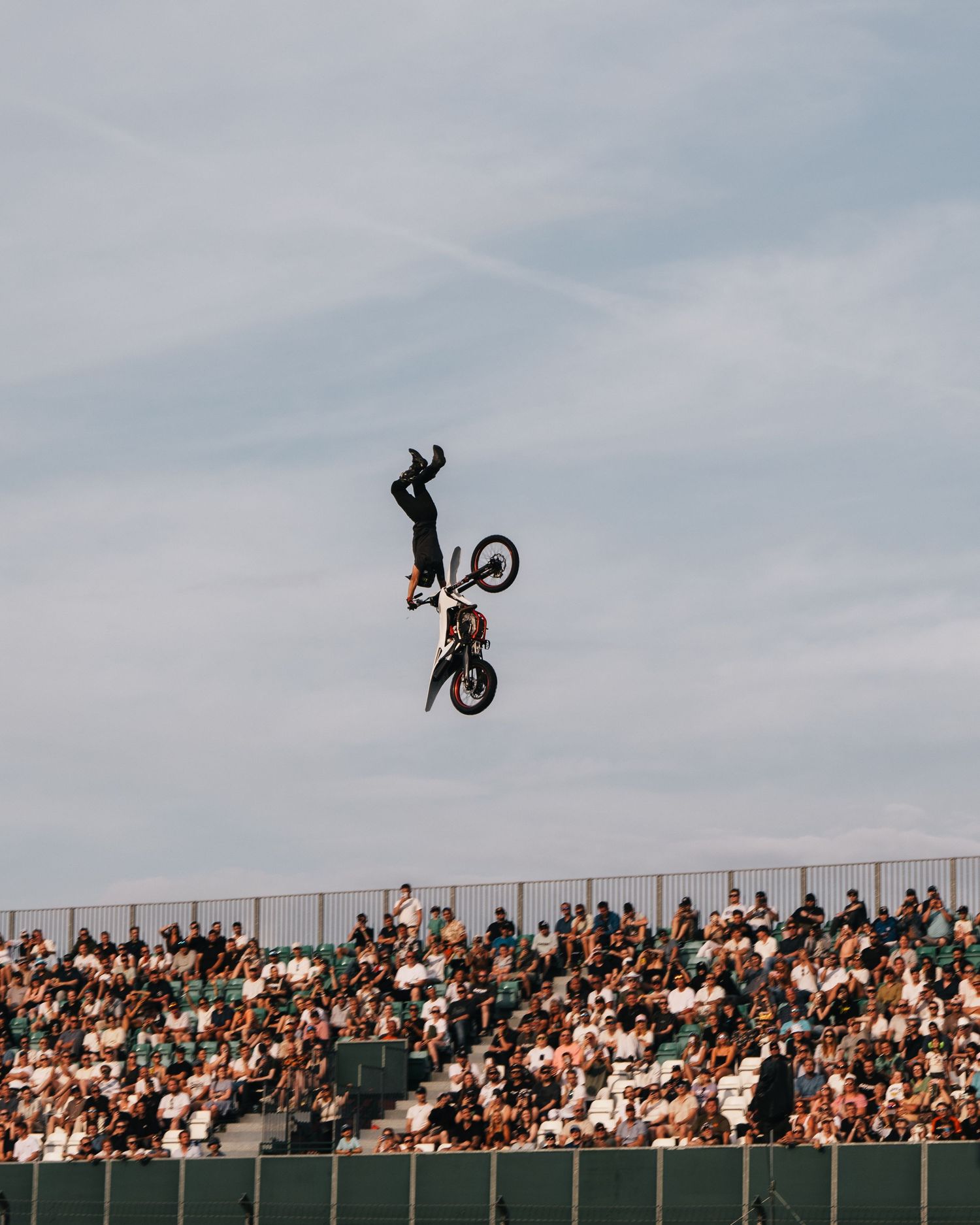 BMX rider performs aerial backflip trick high above ramp with crowded stadium bleachers in background.