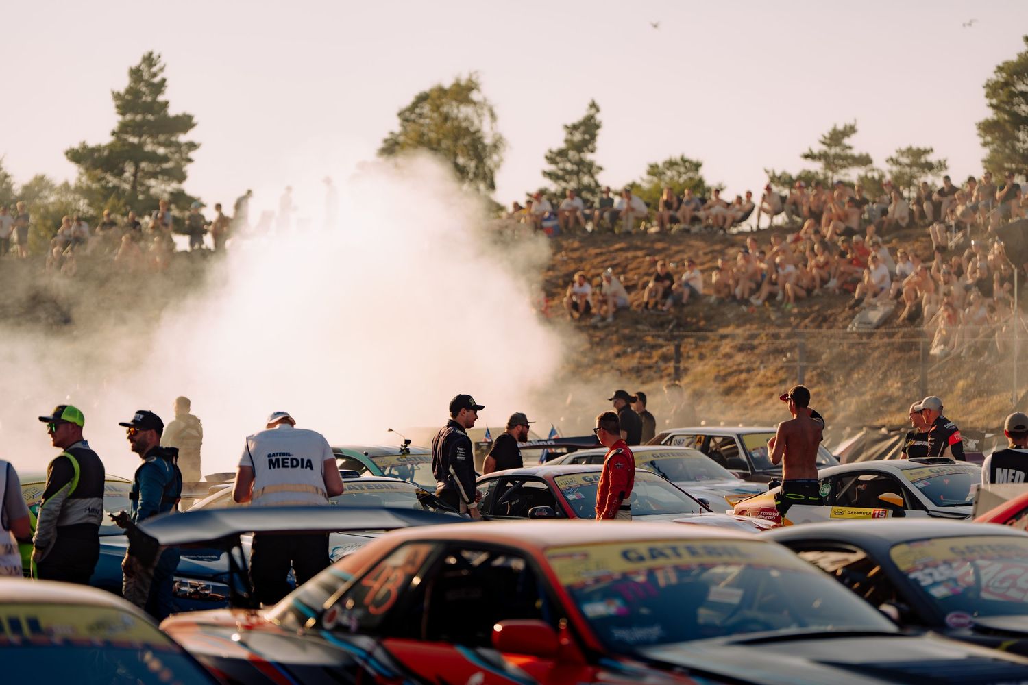 A crowd gathers around cars as burnout smoke fills the air at an outdoor motorsport event.