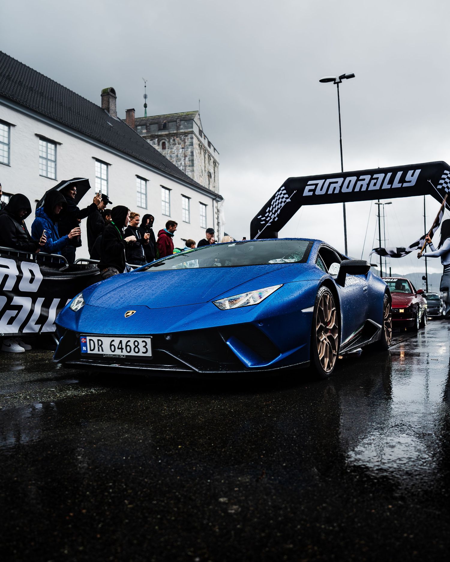 A sleek blue supercar parked in wet conditions with Eurorally branding visible in the background.
