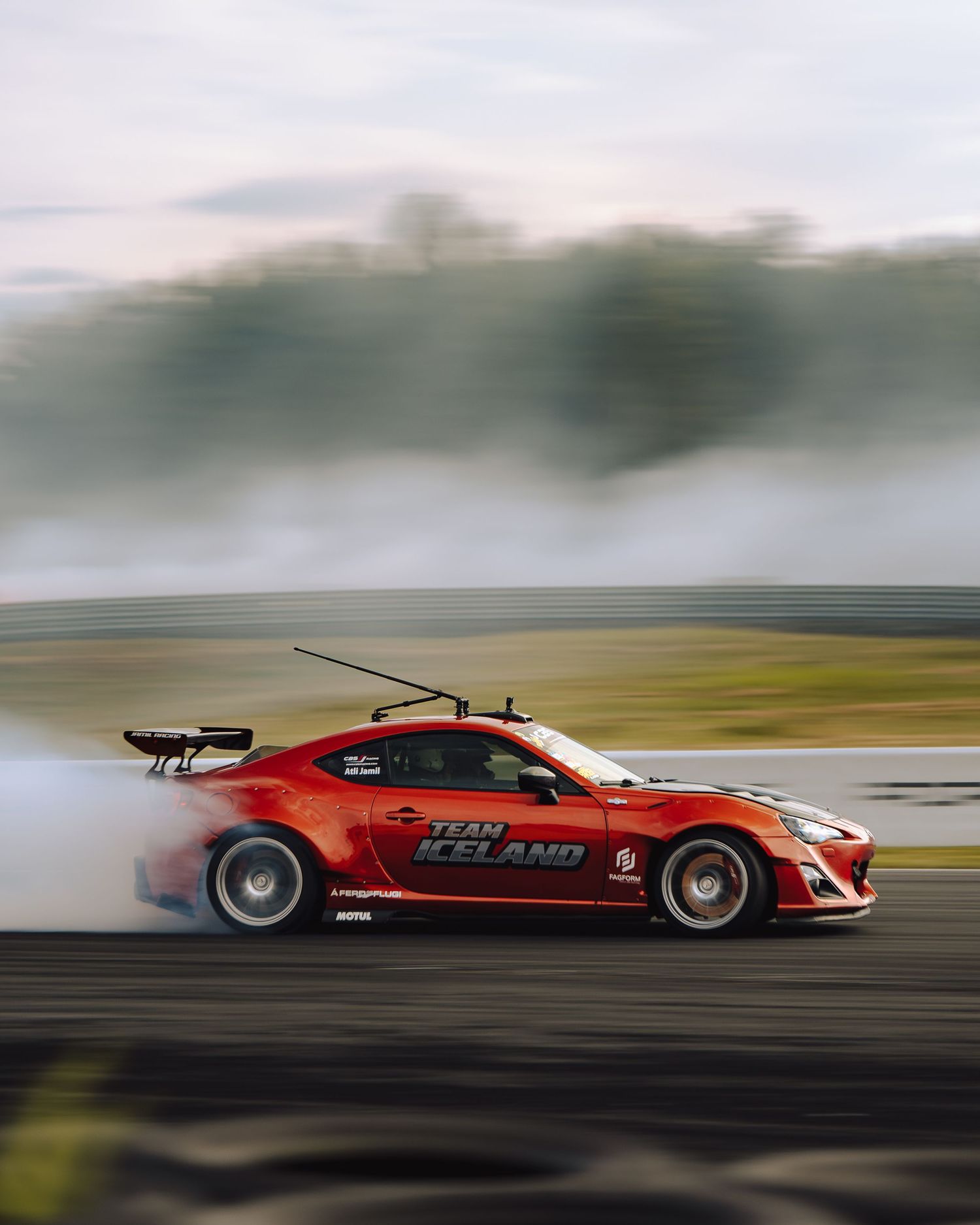 A red sports car speeds along a racetrack creating a dramatic motion blur effect against a cloudy sky.