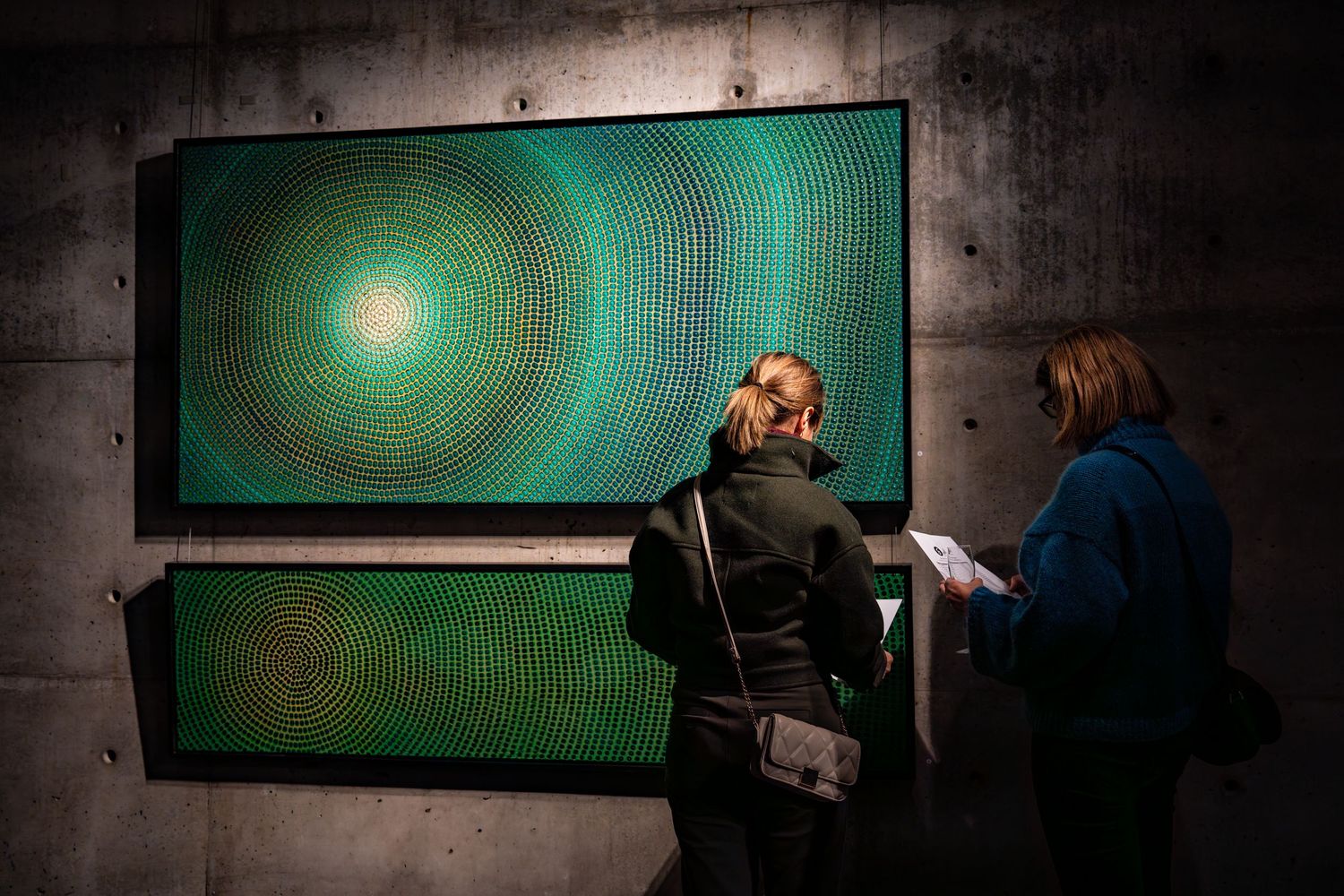 Visitors examine a mesmerizing green spiral artwork mounted on concrete gallery walls.