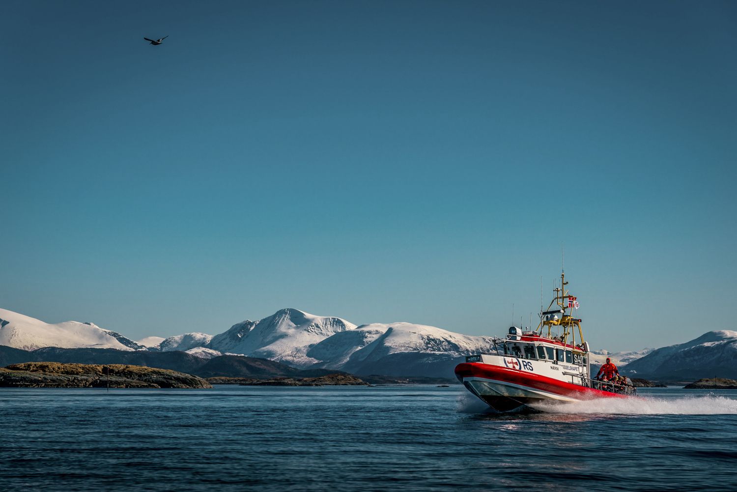 Red and white rescue boat cruising through icy Norwegian waters with snow-capped mountains in the background.