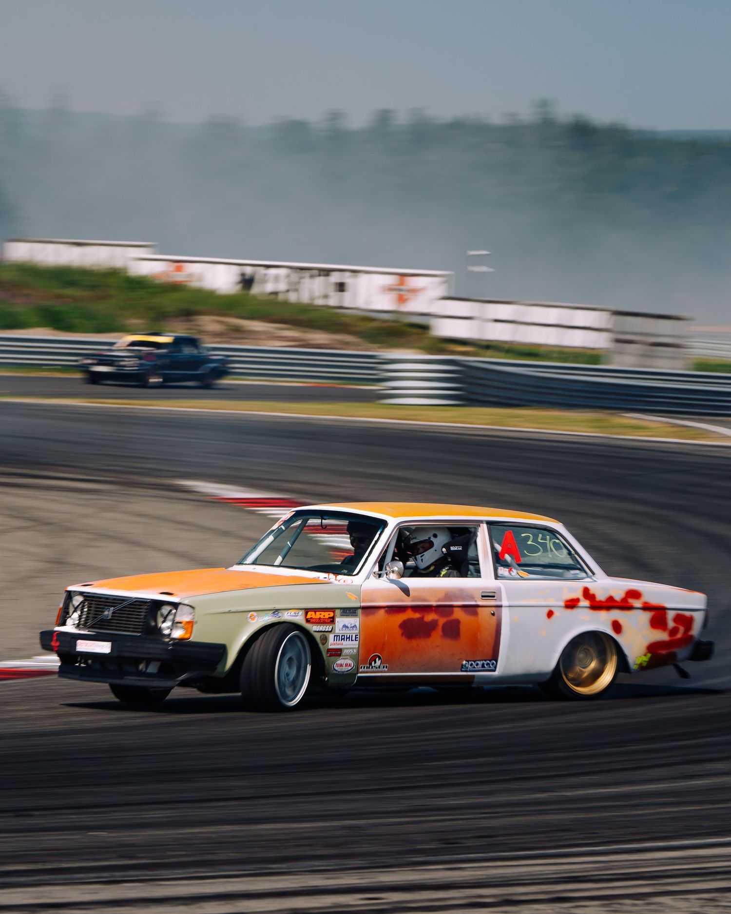 A vintage orange and white Volvo race car drifts around a racetrack corner during a motorsport competition.