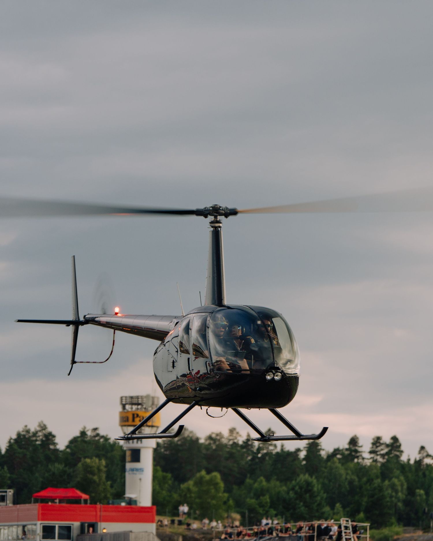 A black Robinson R44 helicopter hovers low over a grassy field with trees in the background during an overcast day.