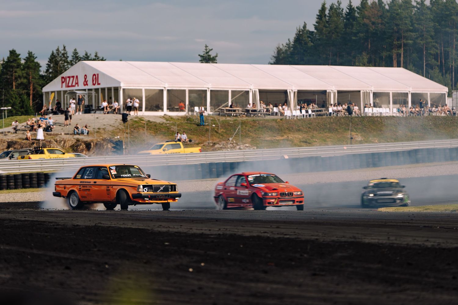 Multiple cars race and drift around a track with tire smoke and evergreen trees in the background.