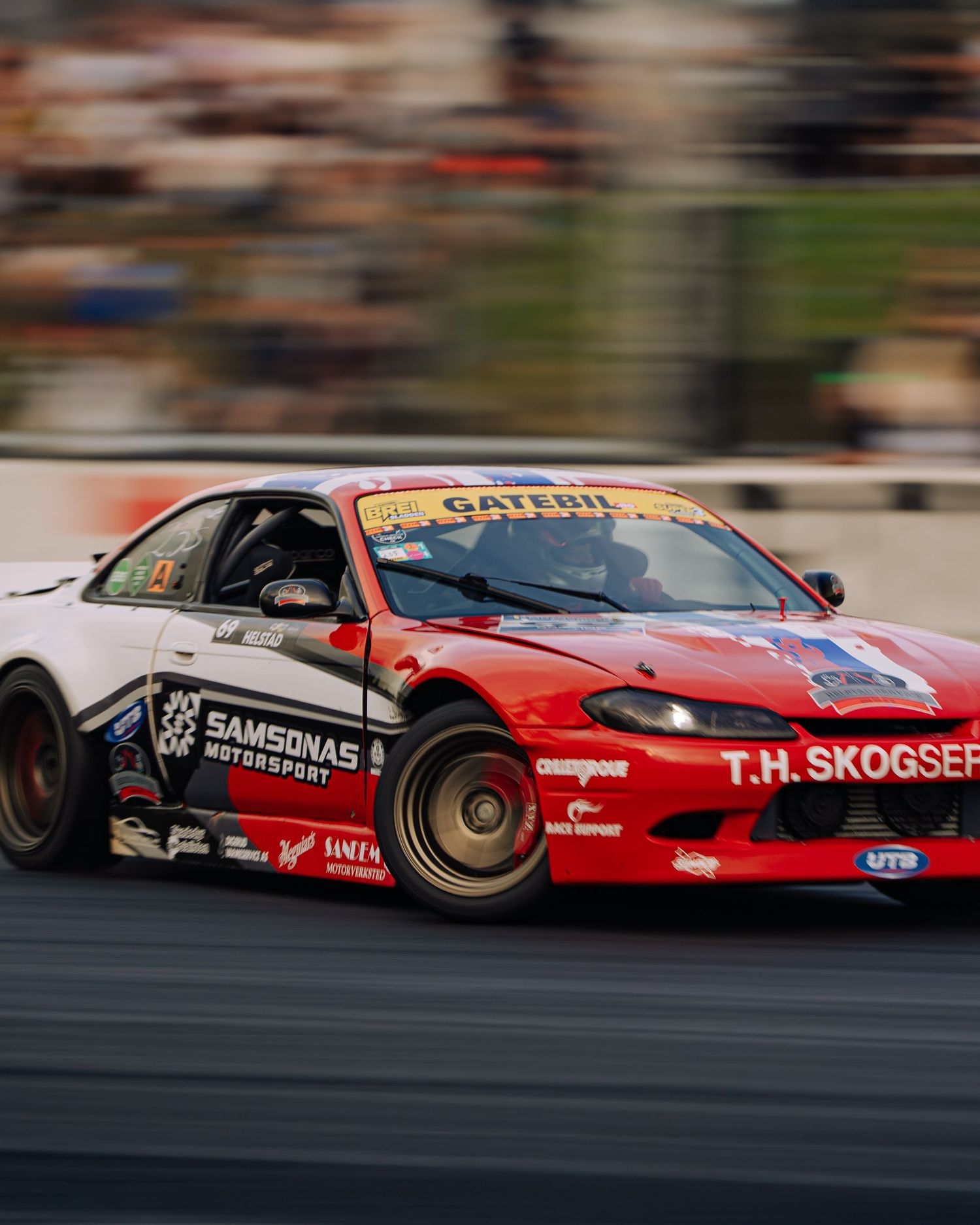 A red drift car with racing livery slides sideways on a racetrack during a competition event.