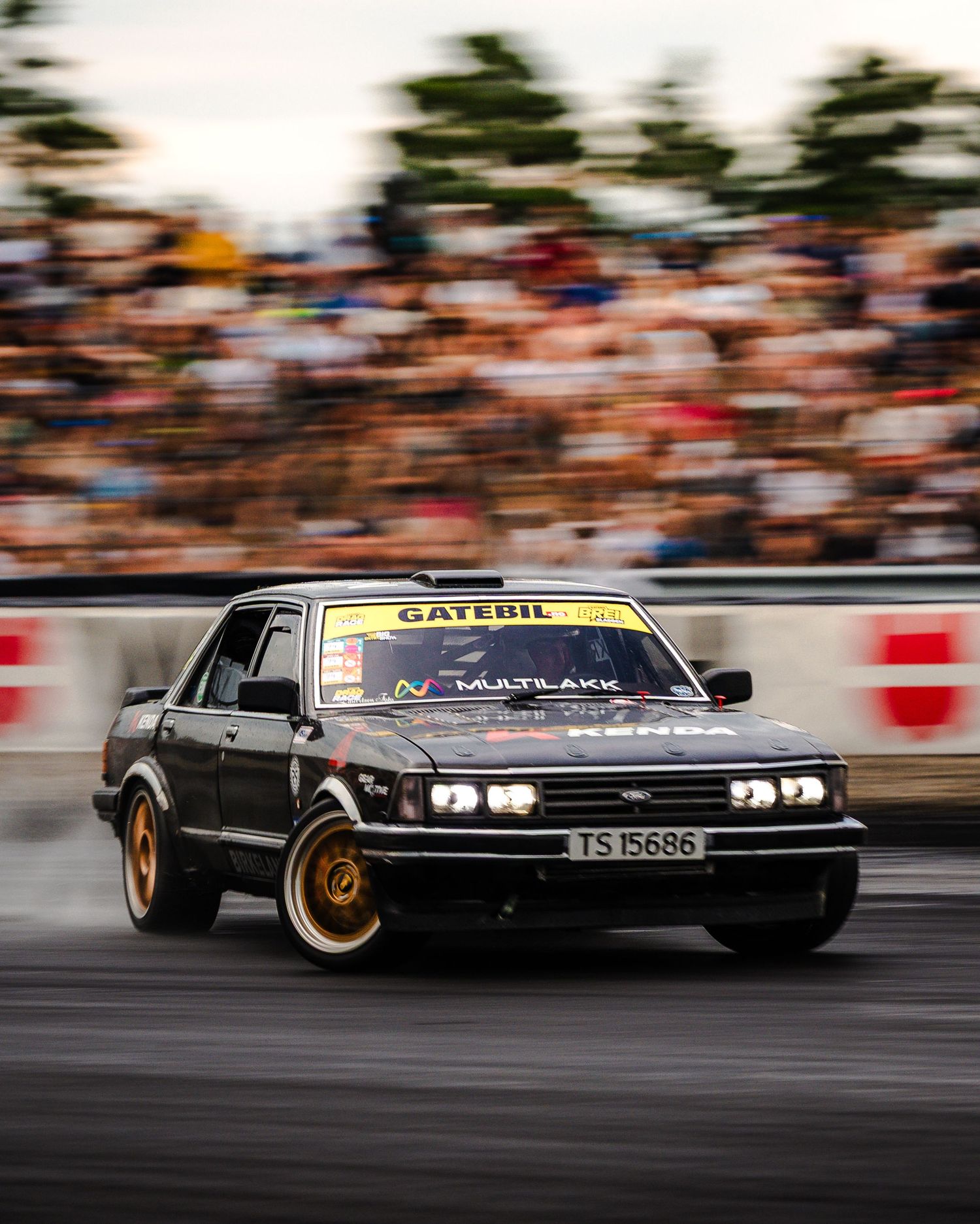 Black vintage car drifting on a racetrack with motion blur and crowd in background.
