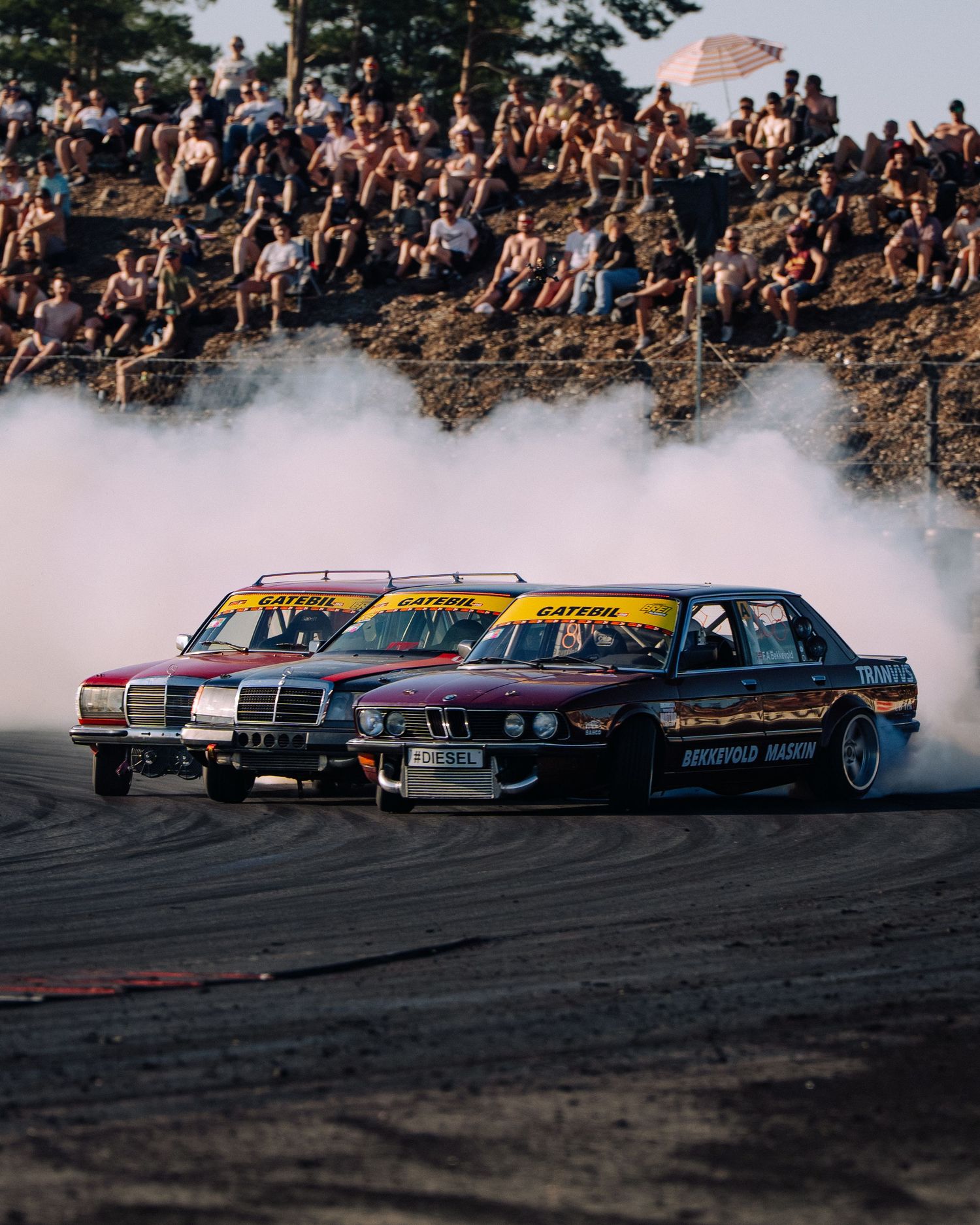 Multiple drift cars perform synchronized drifting on a track with a large crowd watching from elevated rocks in the background.