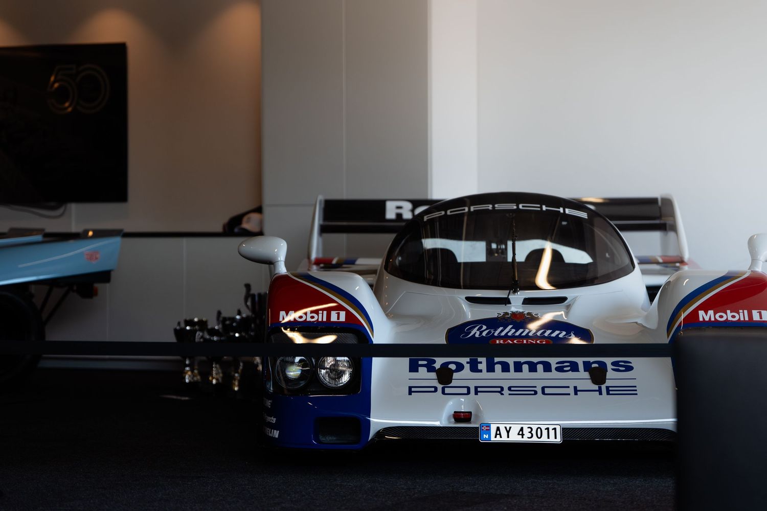 Vintage Rothmans racing car model with large rear spoiler and blue and white livery displayed in an indoor setting.