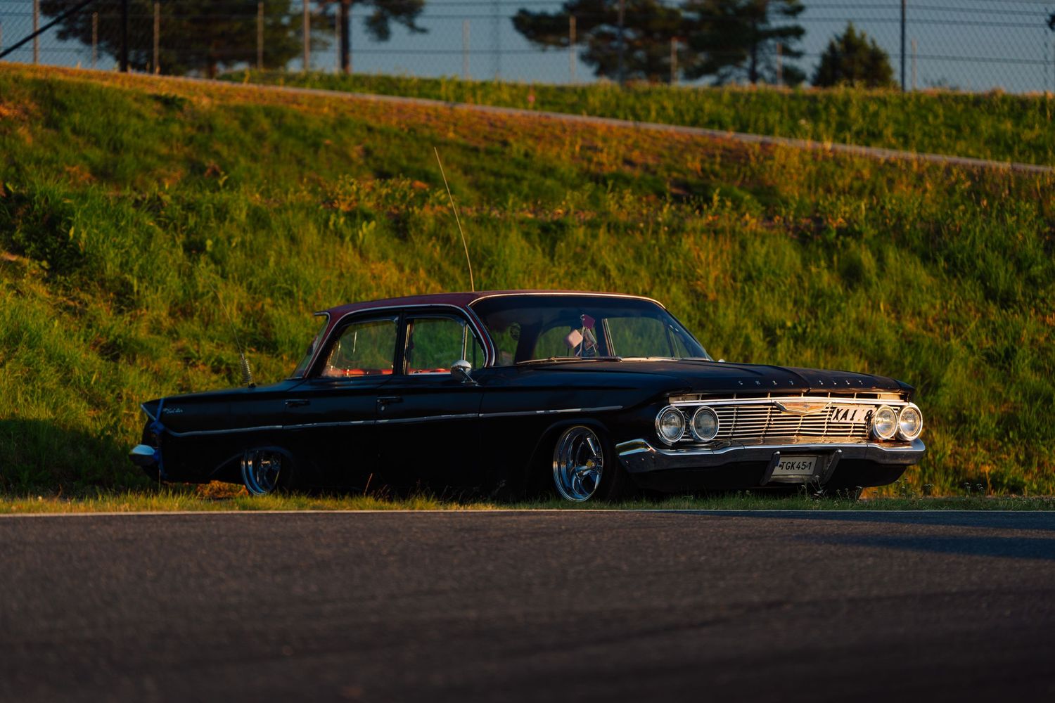 Classic black vintage car silhouetted against a grassy hillside at sunset with power lines visible in the background.