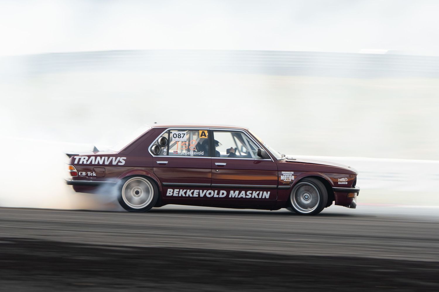 A vintage Cadillac car performs a burnout on a racetrack, creating dramatic smoke from its tires.