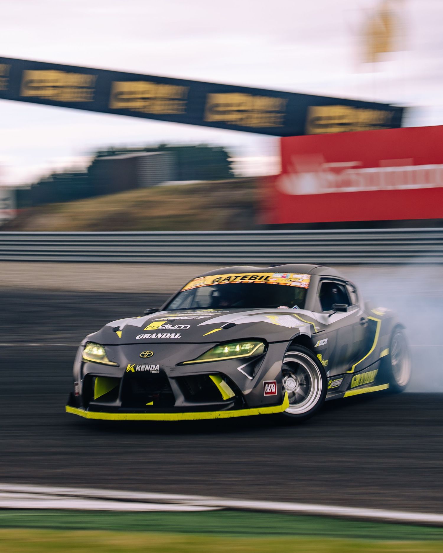 A sports car drifts around a racetrack with smoke trailing from its tires during a competitive drift event.