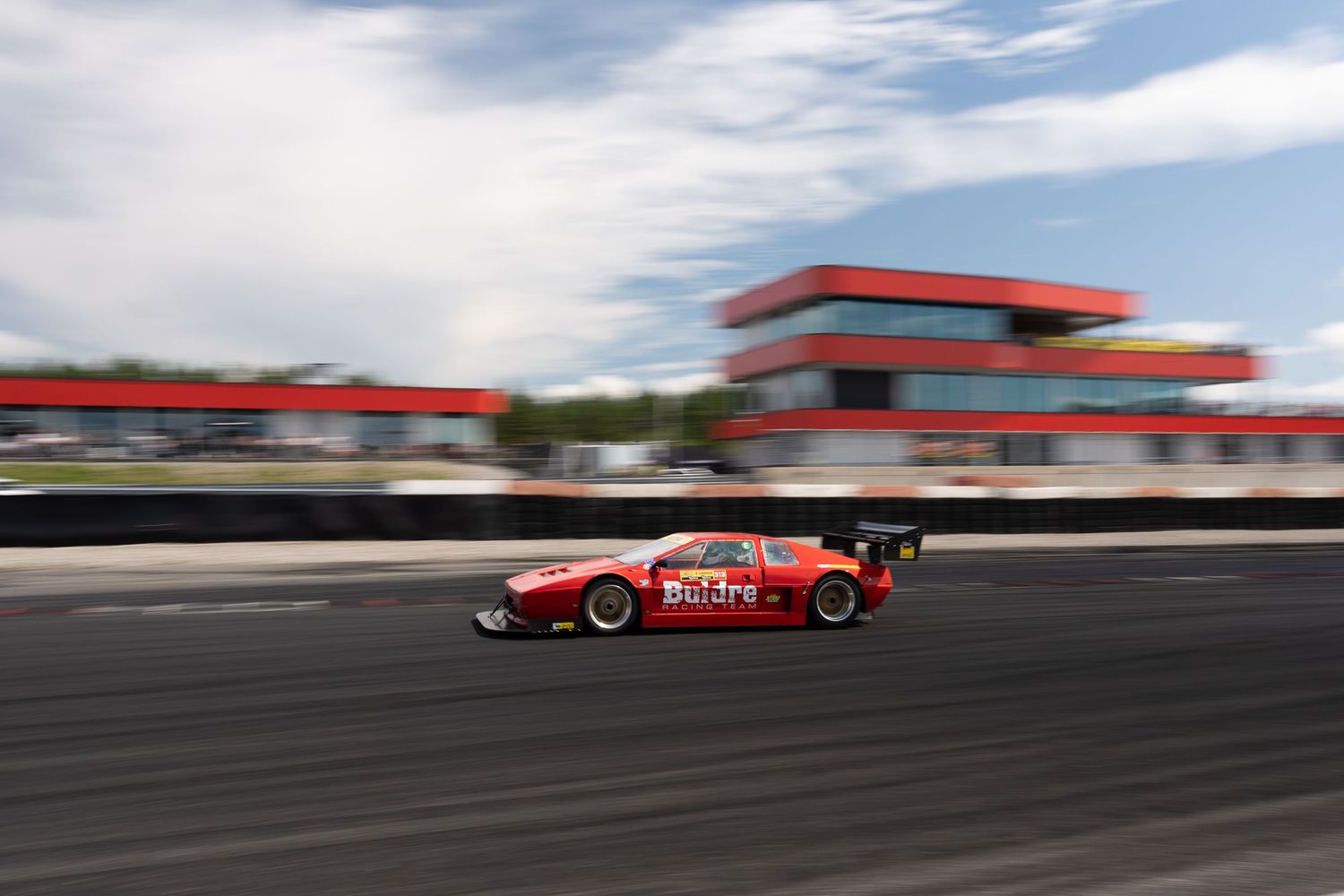 Red race car speeds past blurred background on professional racing track during competitive motorsport event.