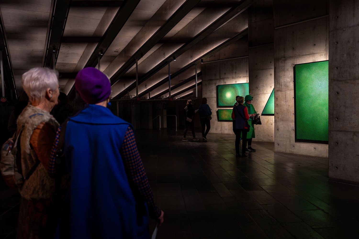 Museum visitors explore a dark gallery space with glowing green artworks mounted on concrete walls.