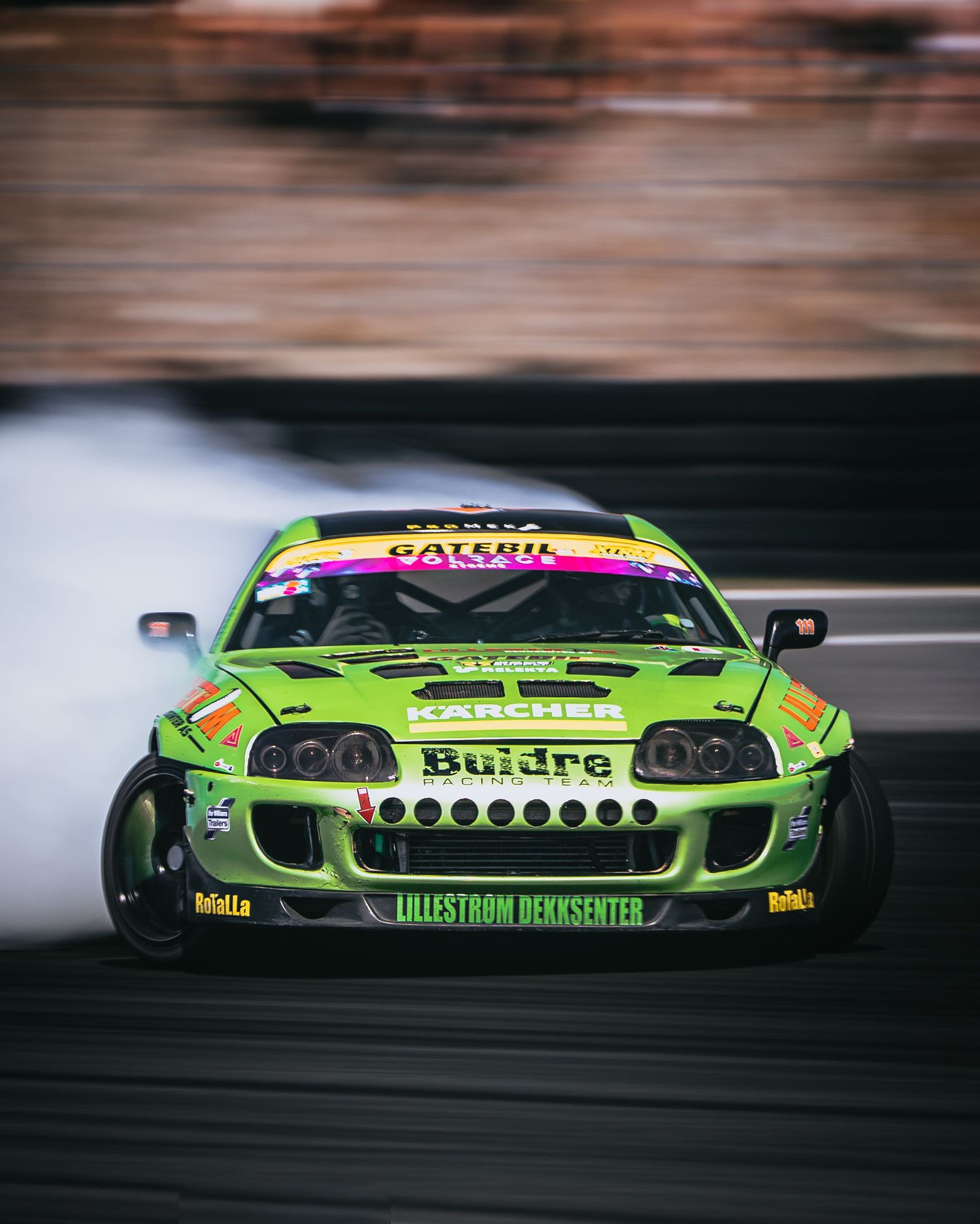 A bright green race car with Buidra branding speeds past in a motion-blurred action shot during a motorsport event.
