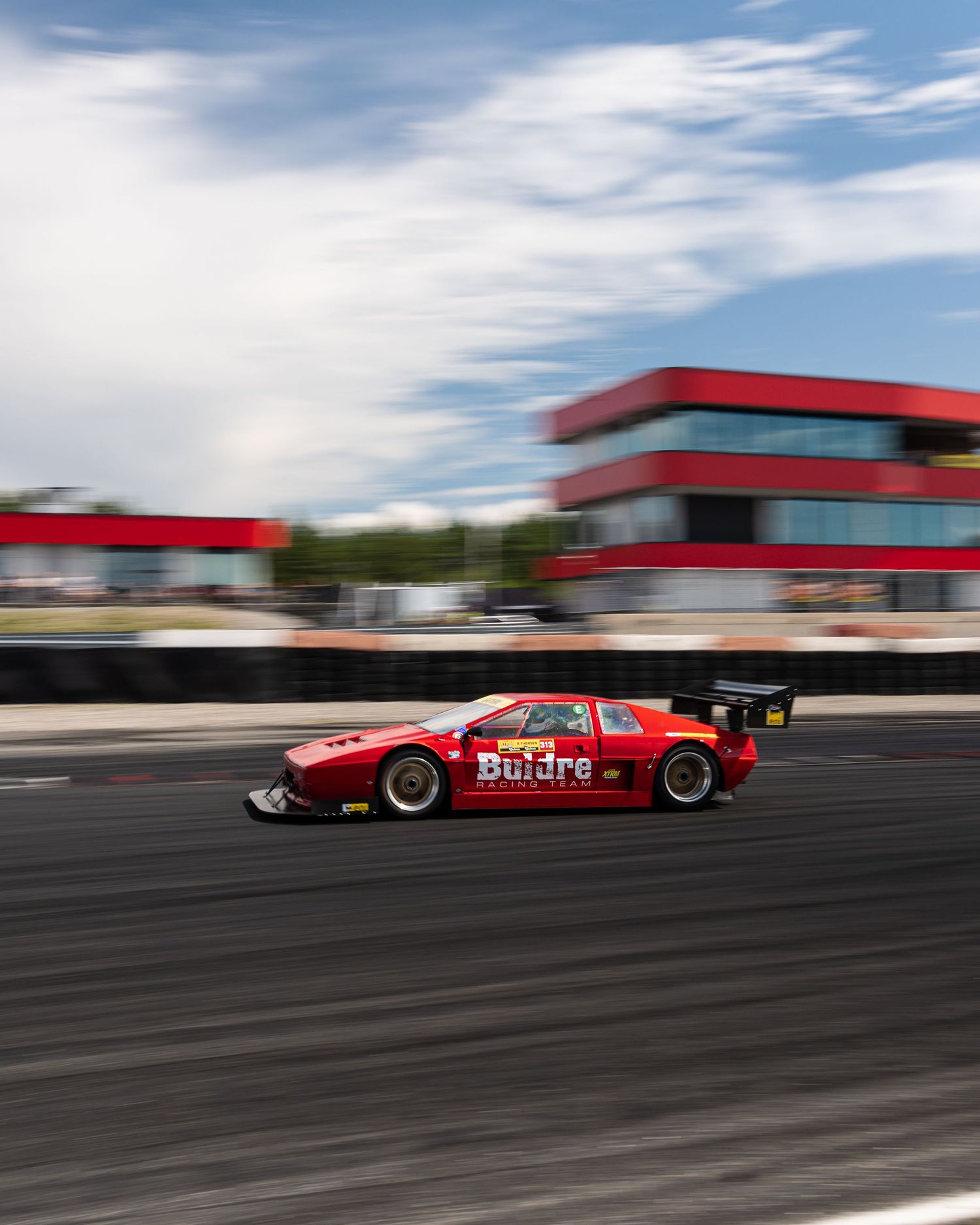 A red race car speeds along a track with motion blur against buildings and cloudy sky in the background.