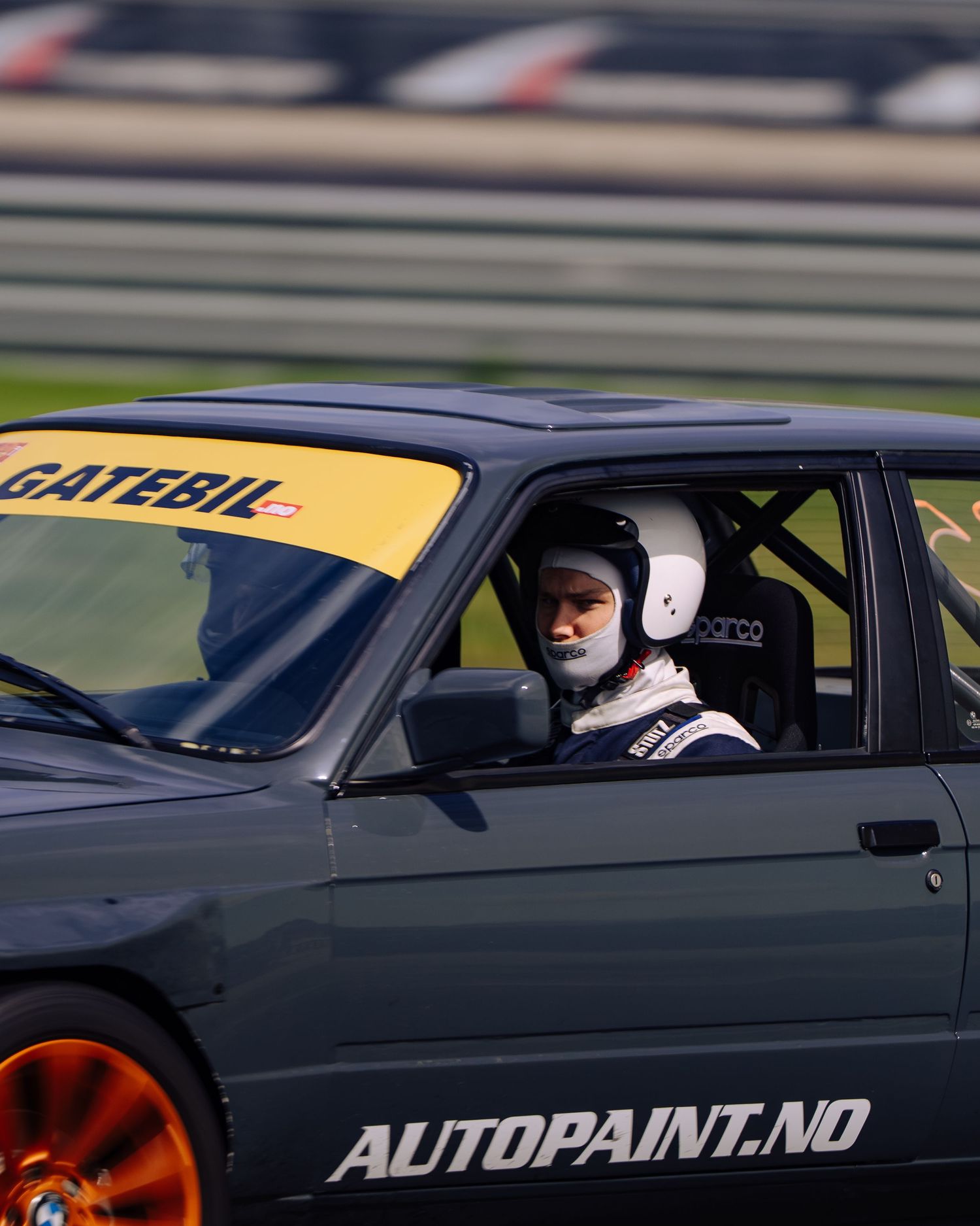 A dark gray race car with orange wheels and Autopaint.no branding sits on a racetrack.