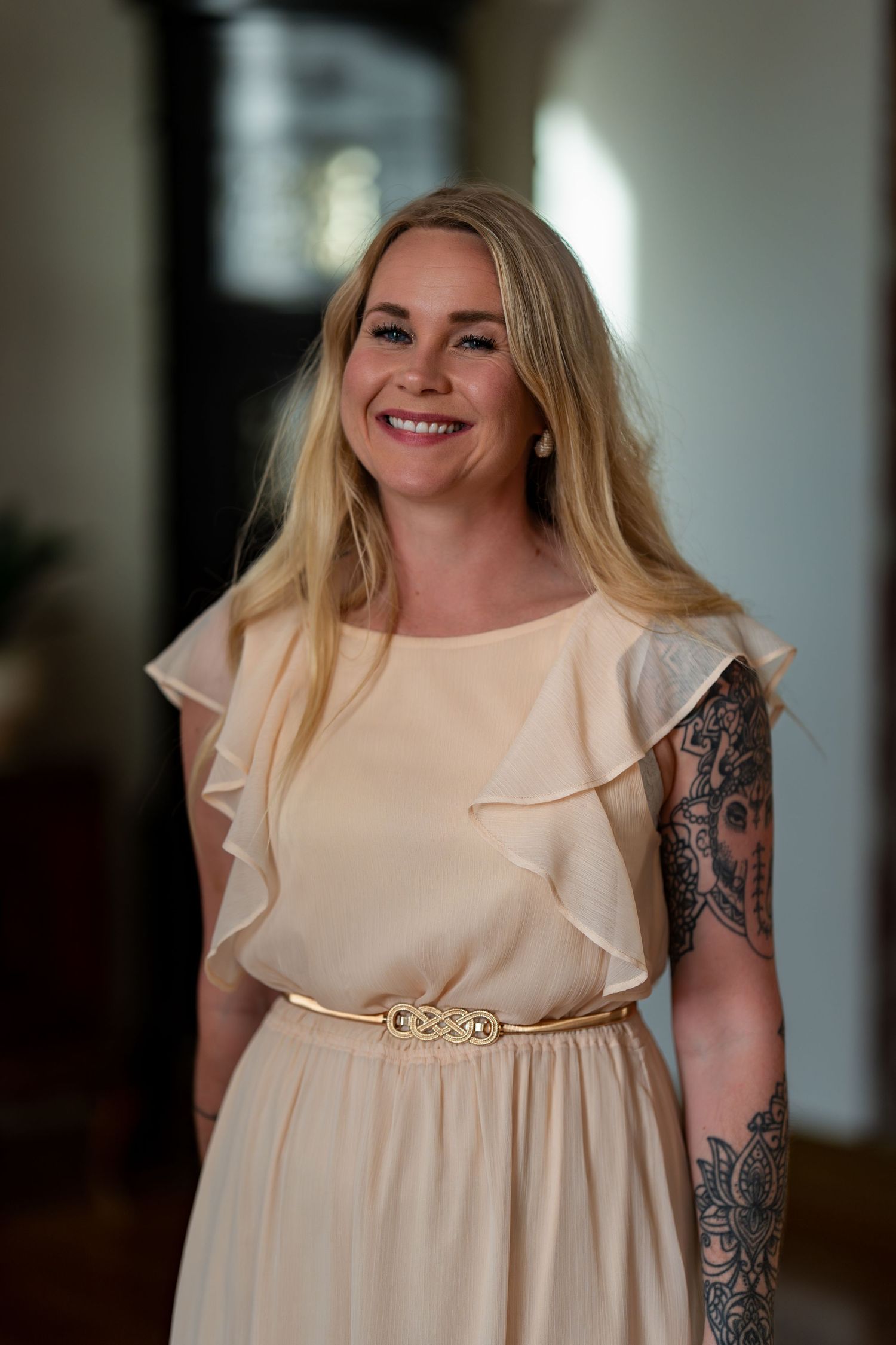 Woman in cream-colored dress with ruffled sleeves and visible tattoos smiling at formal event.